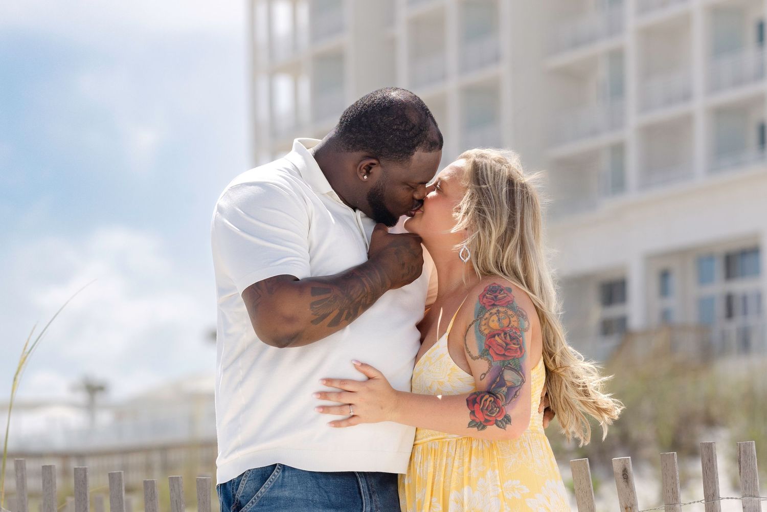 Interracial couple sharing a tender kiss outdoors, woman in yellow dress with floral tattoo sleeve, man in white shirt.