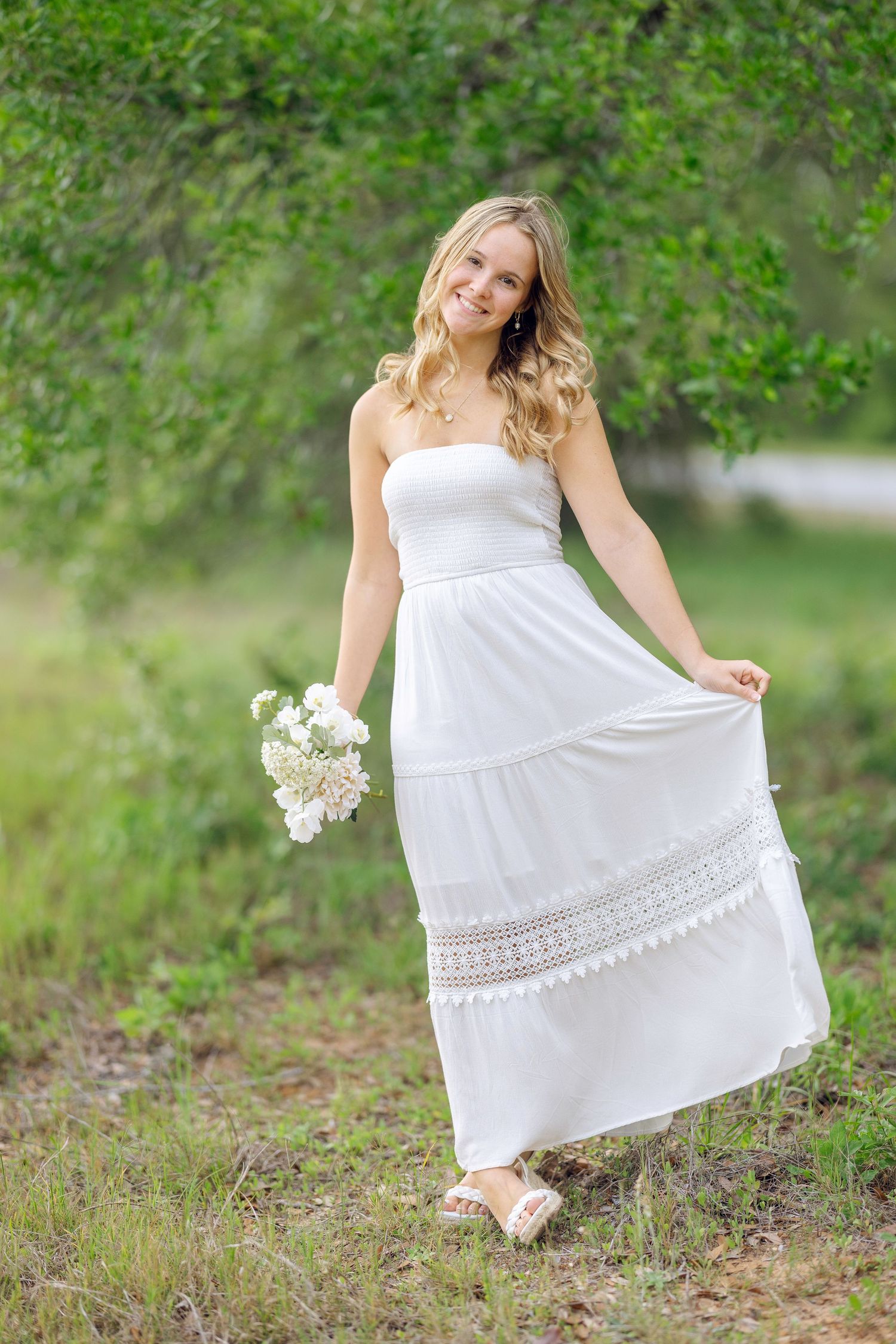 Smiling blonde woman in white strapless dress holding white flowers while standing in nature.