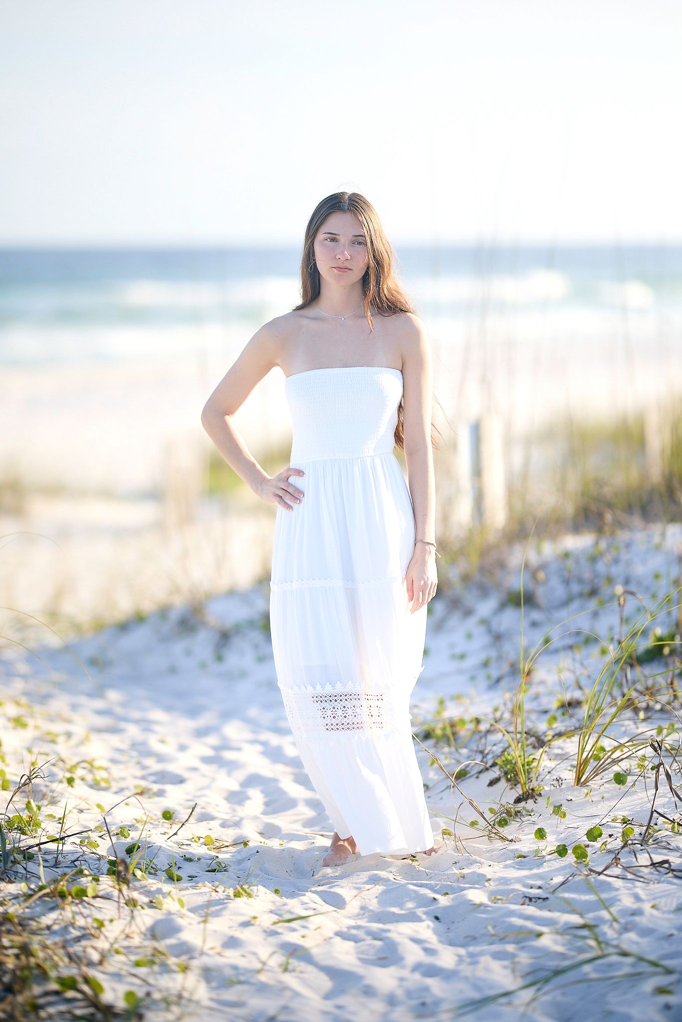 Young woman in white strapless maxi dress standing on sandy beach path with ocean waves in the background.