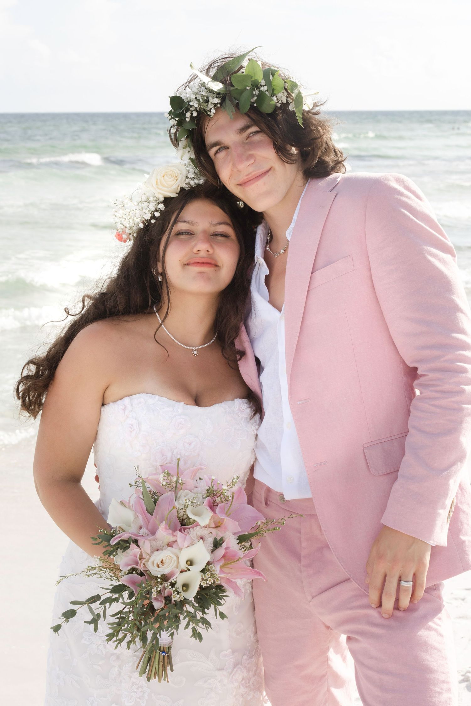 Bride in white dress with floral crown and groom in pink suit pose together on a beach wedding.