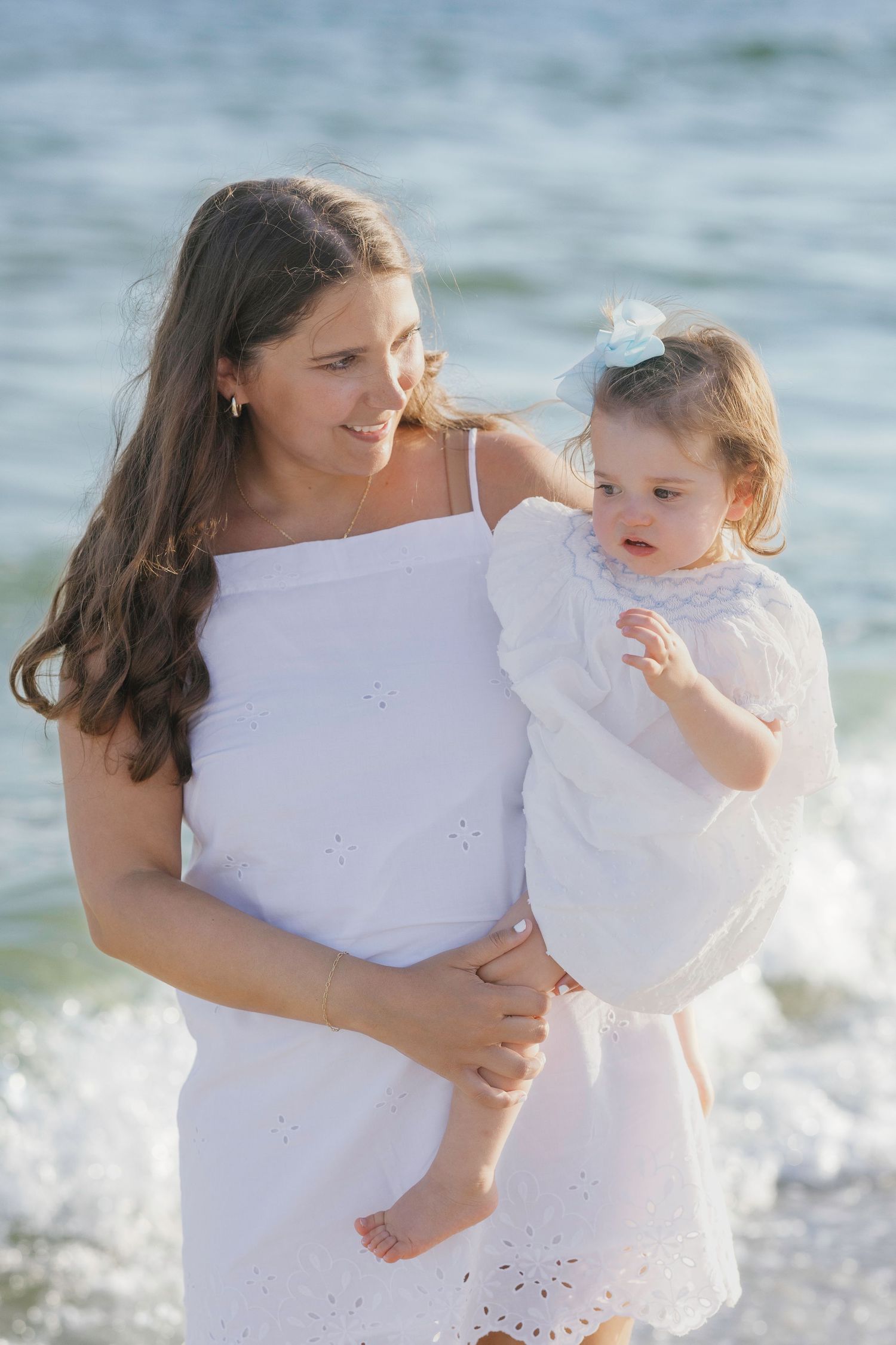 Mother in white dress holding toddler daughter near ocean waves on a sunny beach day.
