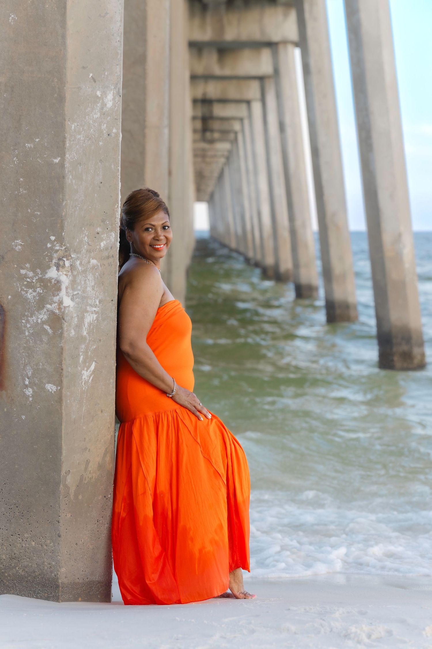 Woman in vibrant orange strapless maxi dress leaning against pier column with ocean waves in background.