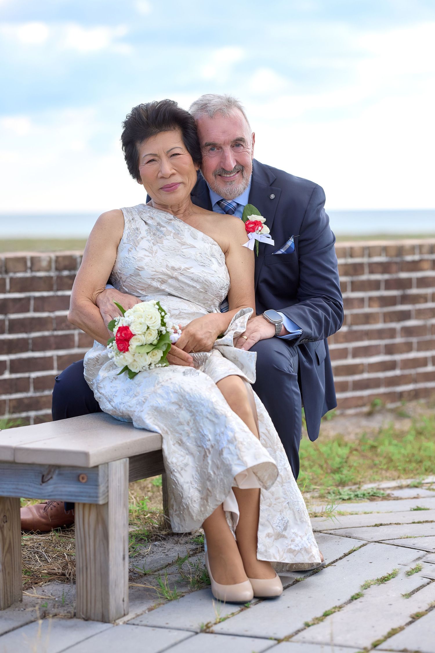Elegant interracial couple in wedding attire posing on a bench outdoors, bride holding bouquet, groom in blue suit.