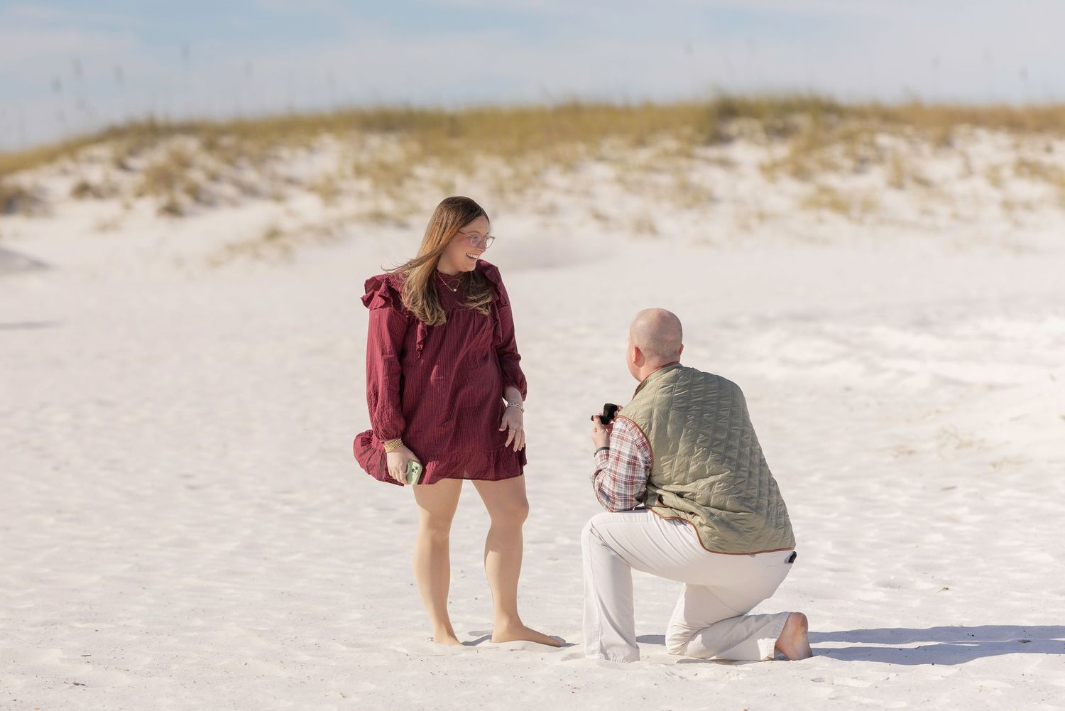 Man kneeling on white sand beach proposing to woman in red jacket during a romantic beach marriage proposal.