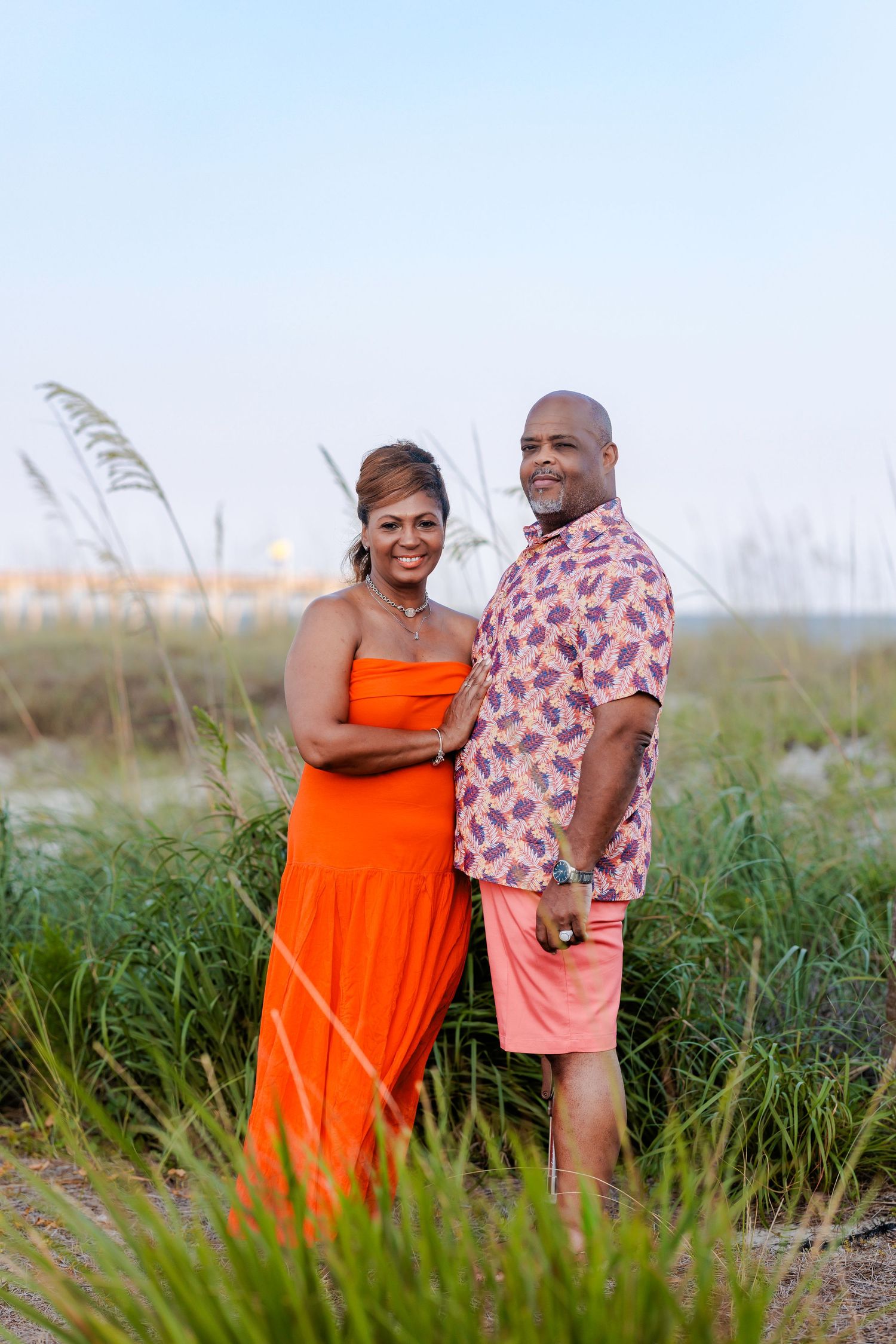 Couple posing together on a beach, woman in orange dress, man in floral shirt and pink shorts, green sea oats background.