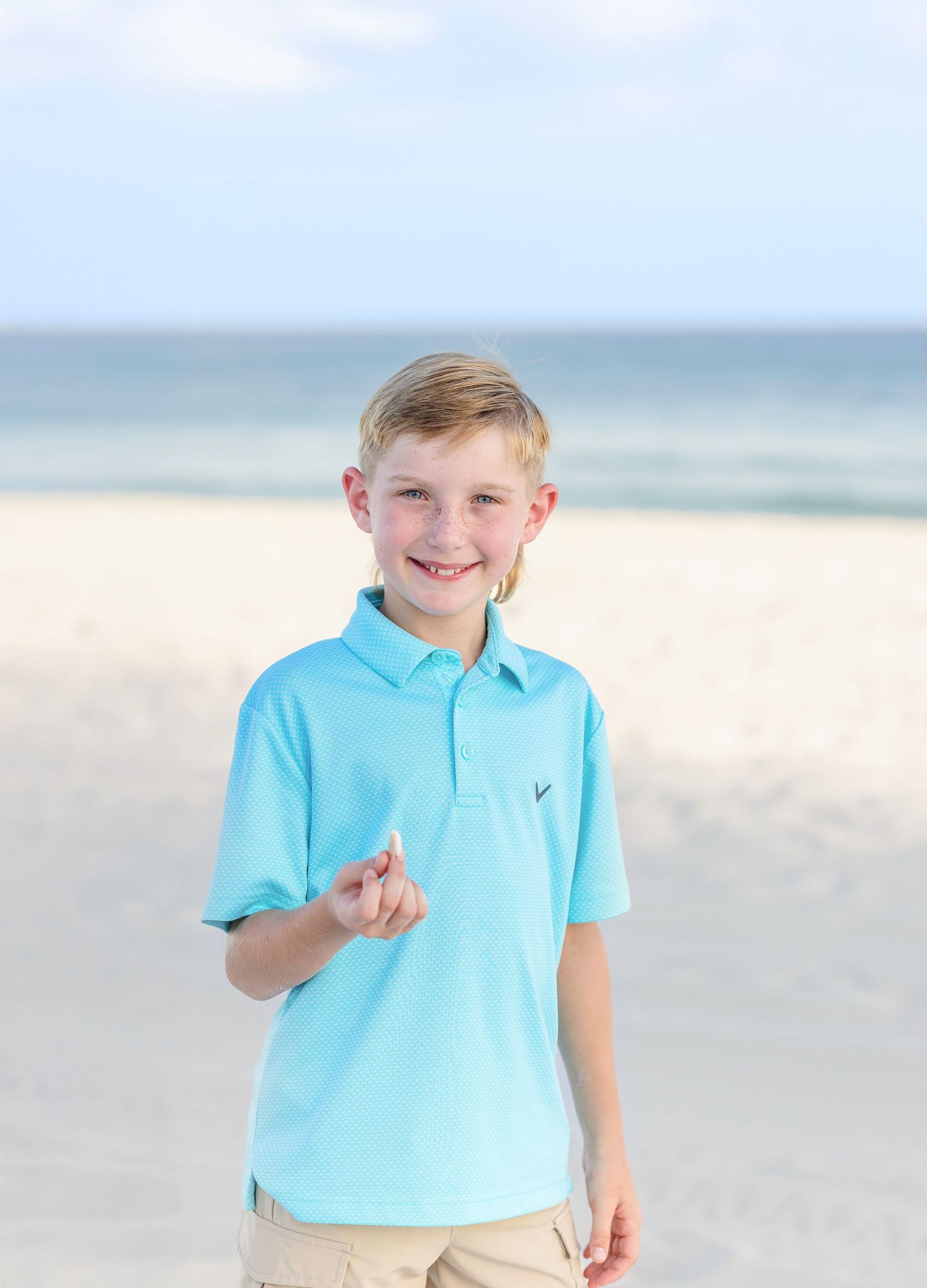 A young person in a light blue polo shirt and khaki shorts smiles while playing on a white sandy beach.