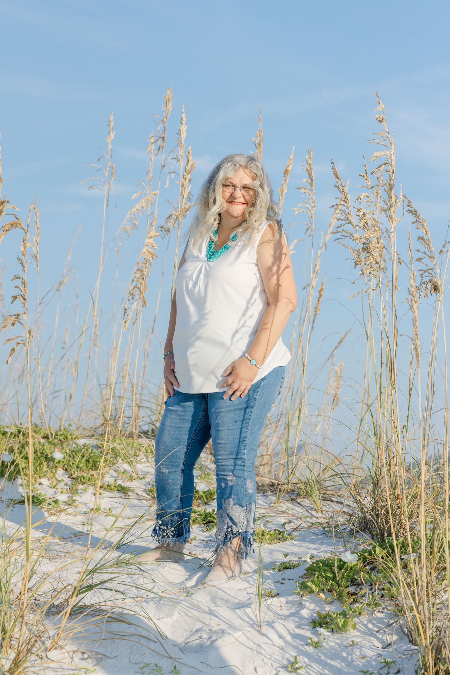 Person standing in white top and jeans among sea oats on a sunny beach with blue skies.