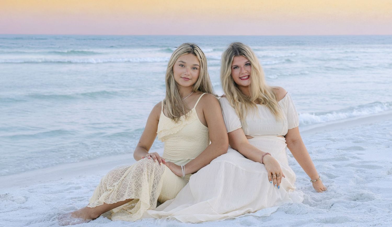 Two people in white dresses sit together on a beach at sunset with ocean waves in the background.