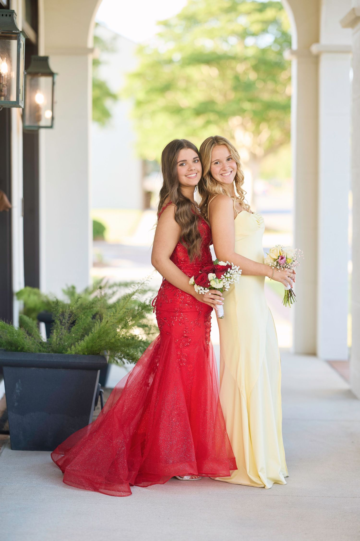 Two young women in formal gowns - one in red and one in yellow - pose together under an outdoor archway.