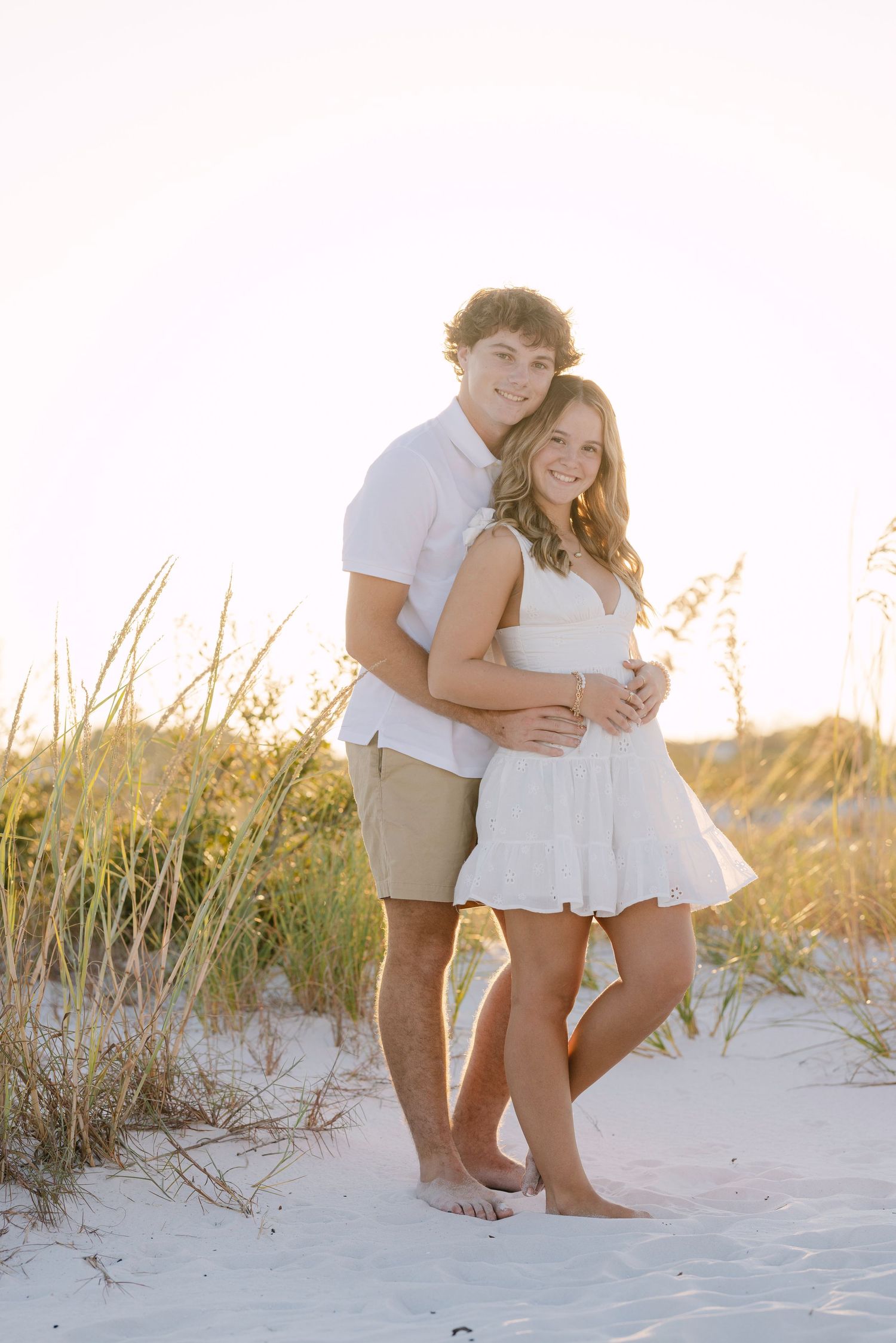A romantic couple embraces and plays together on a sandy beach with beach grass at golden hour sunset.