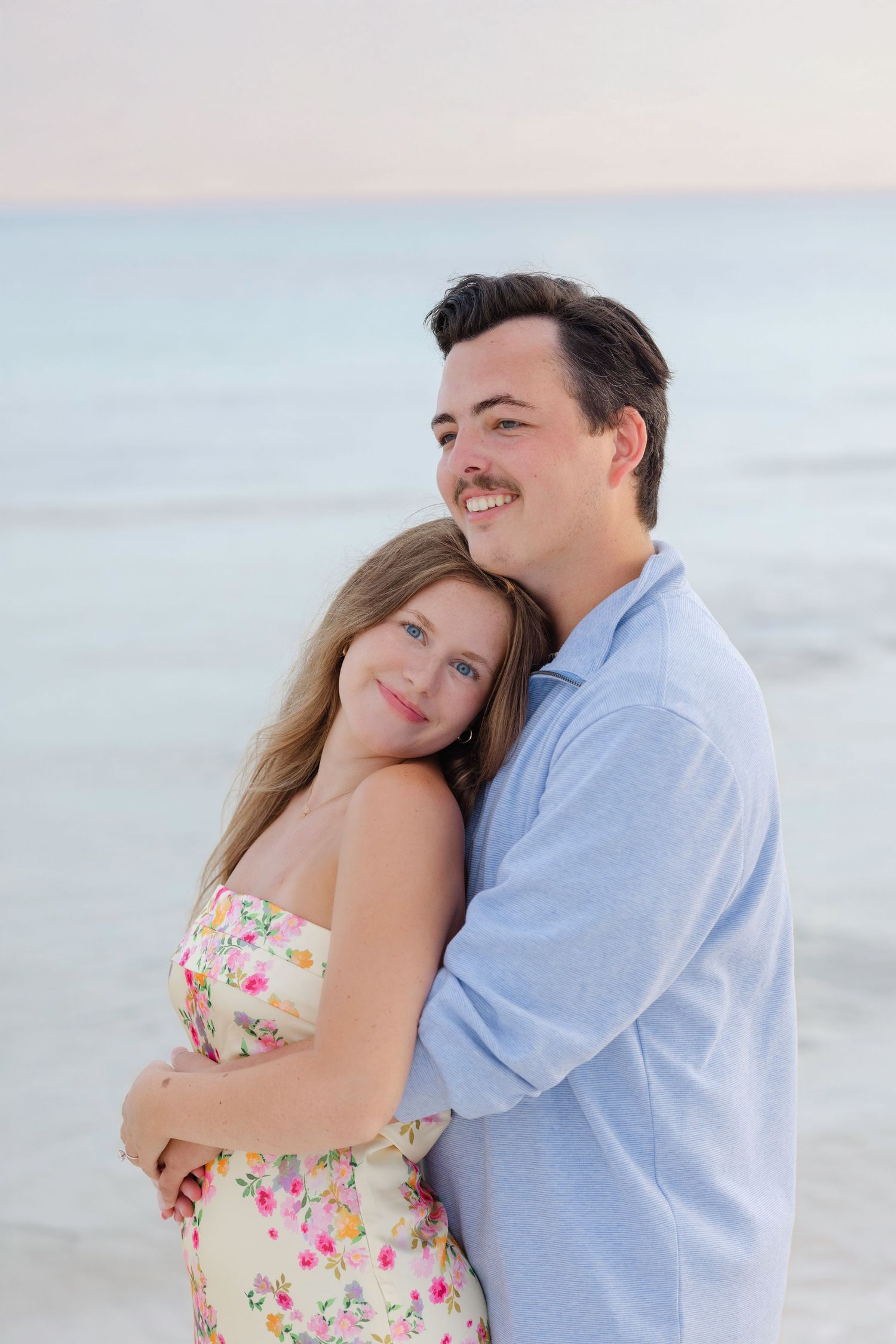 A couple embraces on a beach at sunset, with her in a floral dress and him in a light blue shirt.