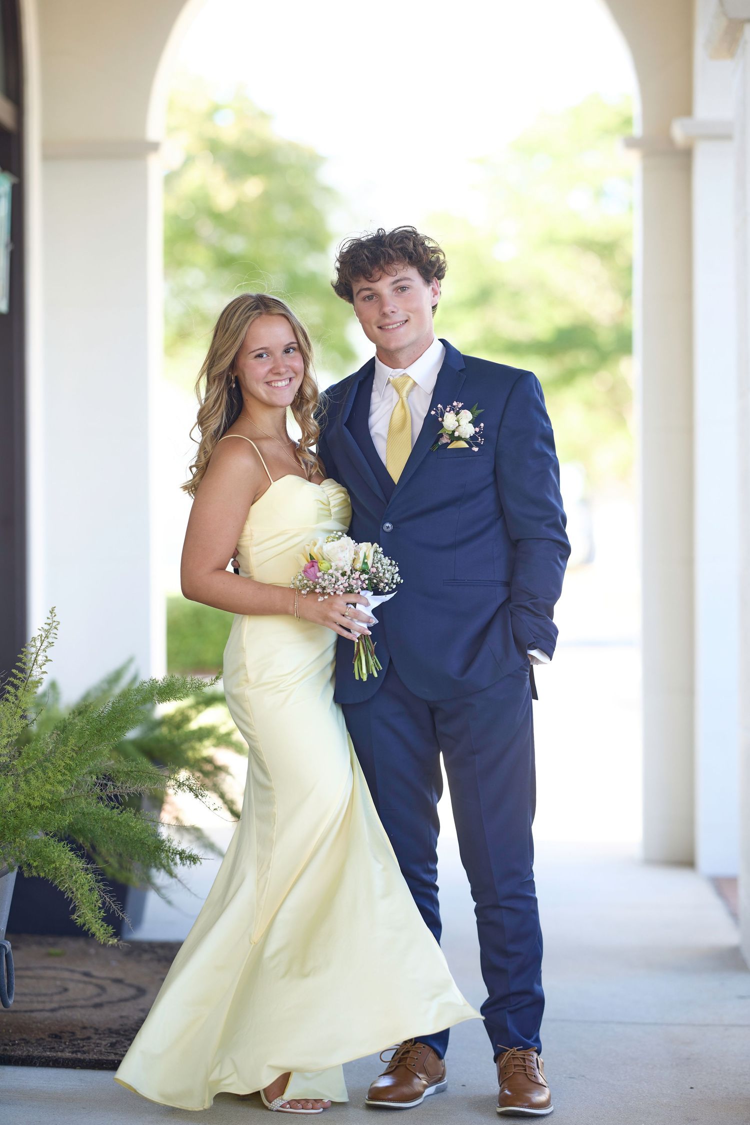 A couple in formal attire poses under an archway, with the woman in a yellow dress holding a bouquet.