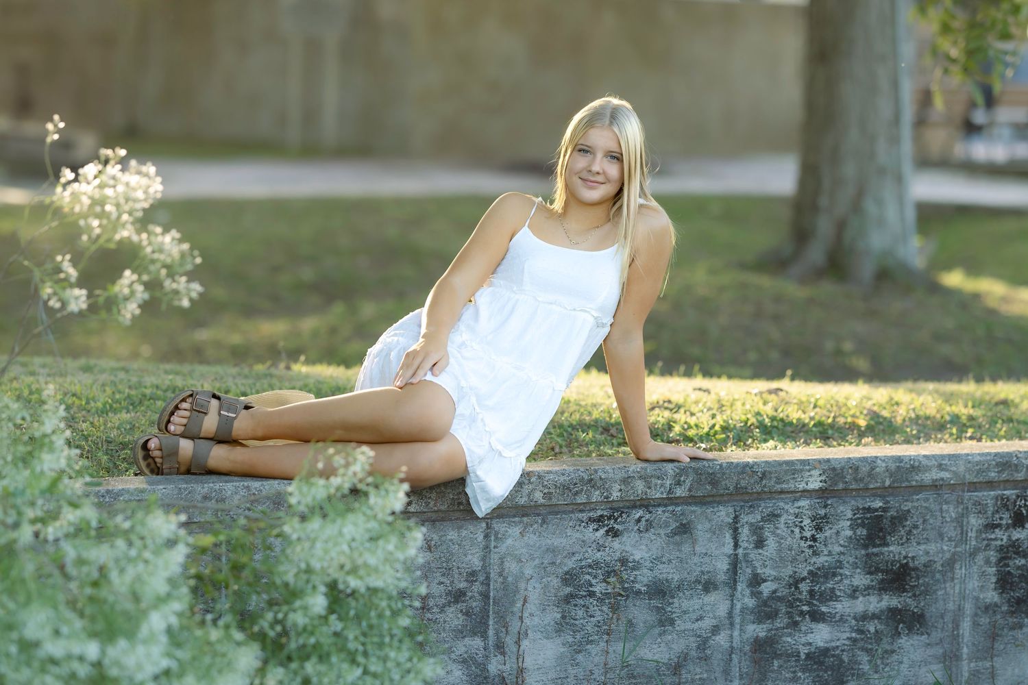 Young blonde woman in white dress sitting on stone wall in sunny park with wildflowers.