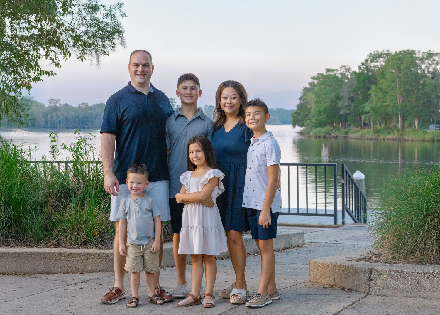 Family of six posing outdoors near a serene lake with lush green trees at dusk.