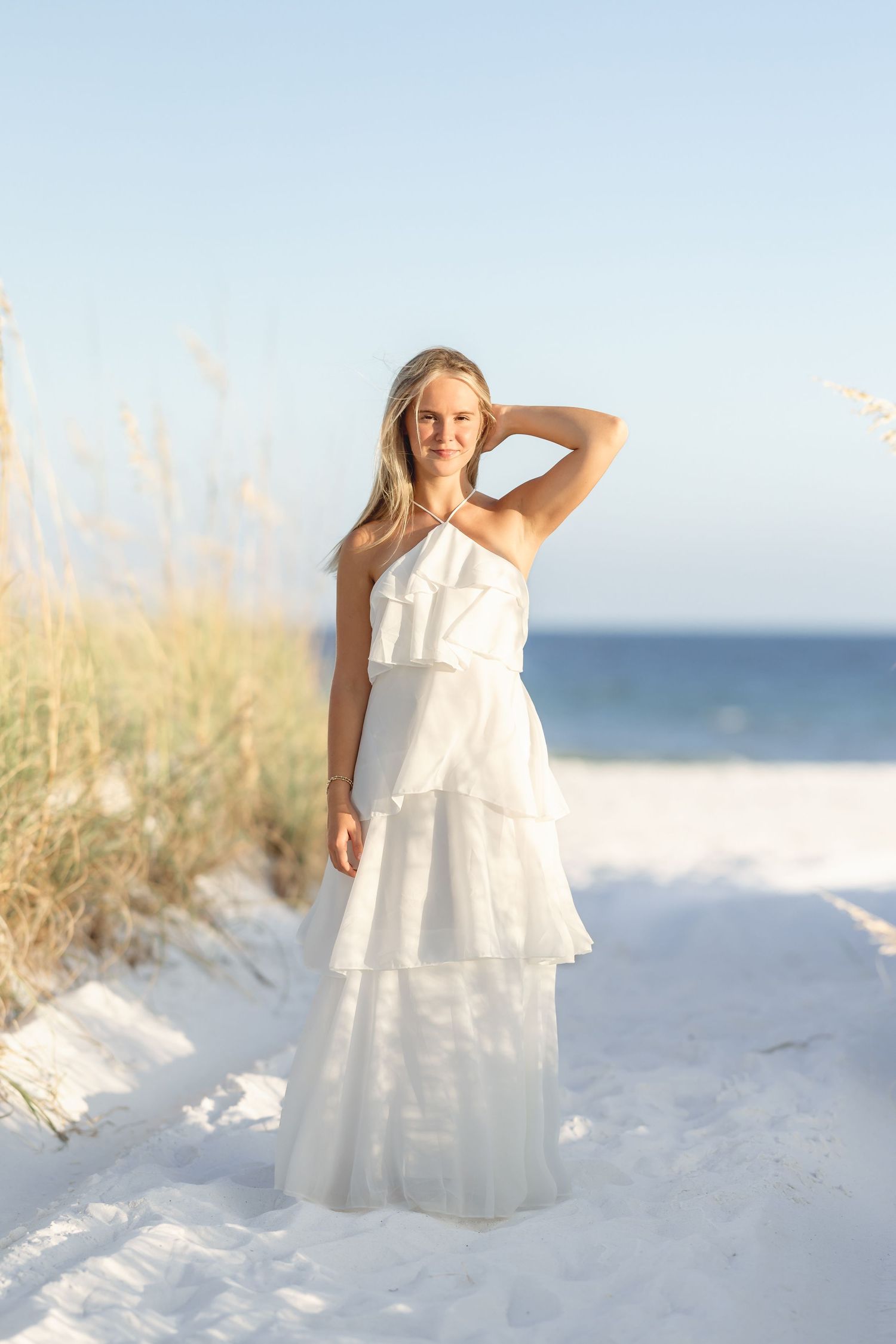 Young blonde woman in white tiered halter maxi dress stands on sandy beach path with sea oats and ocean in background.