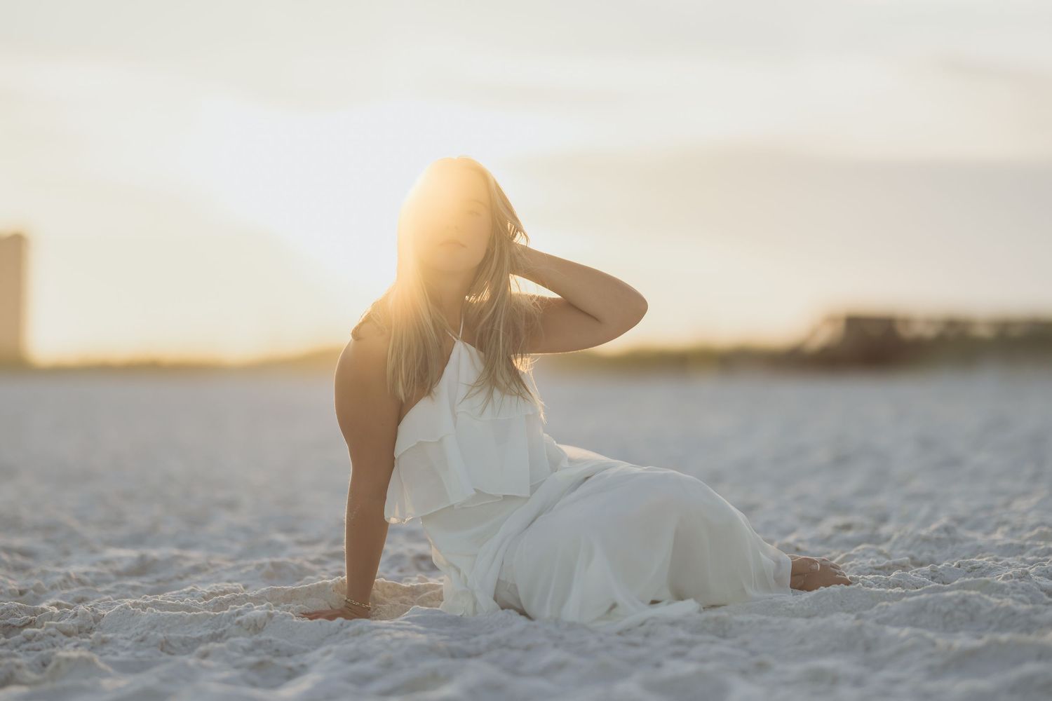 Woman in white dress sitting on sandy beach at golden sunset, hand raised to head in serene pose.
