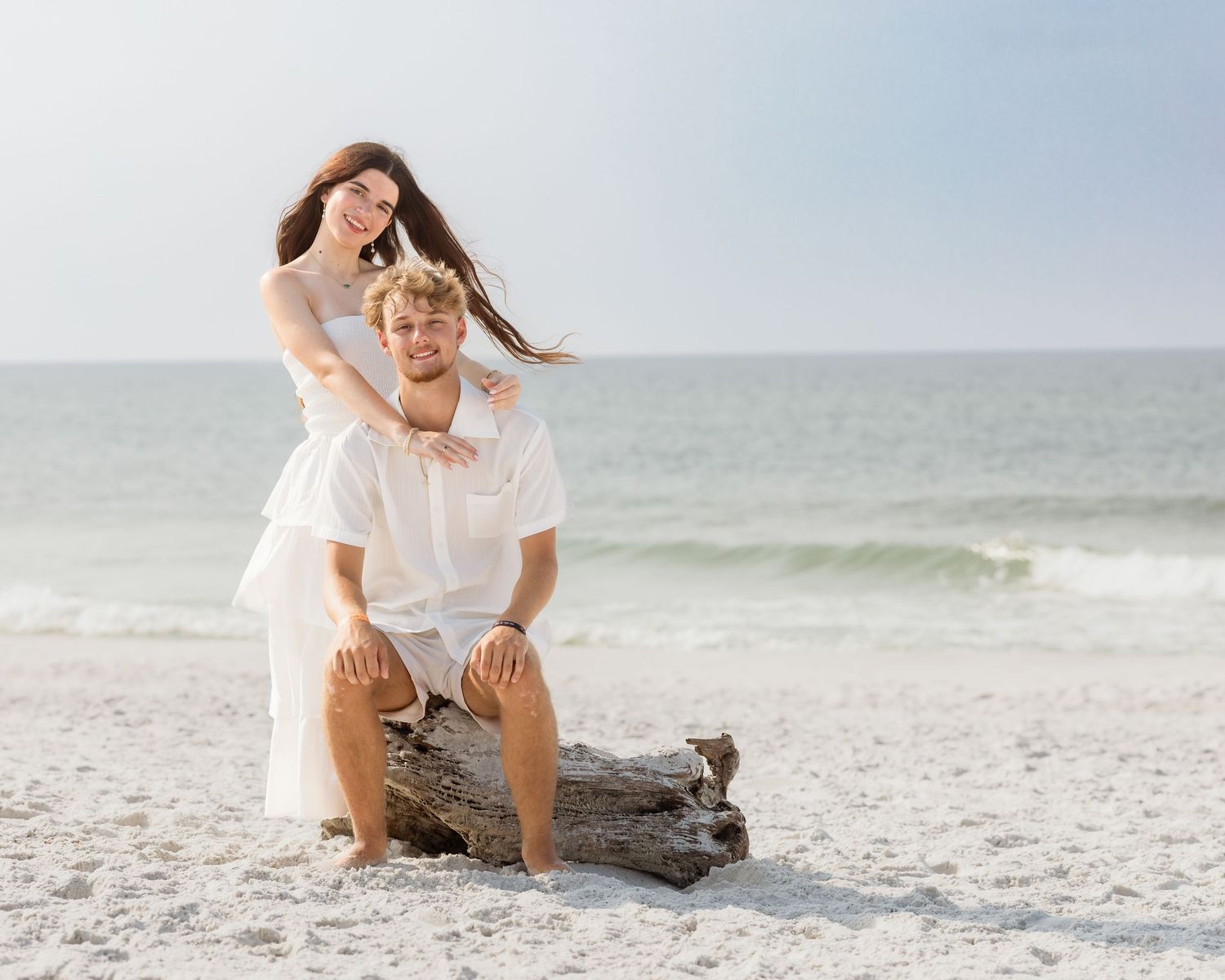 Romantic couple in white outfits posing on driftwood with ocean backdrop on sandy beach.