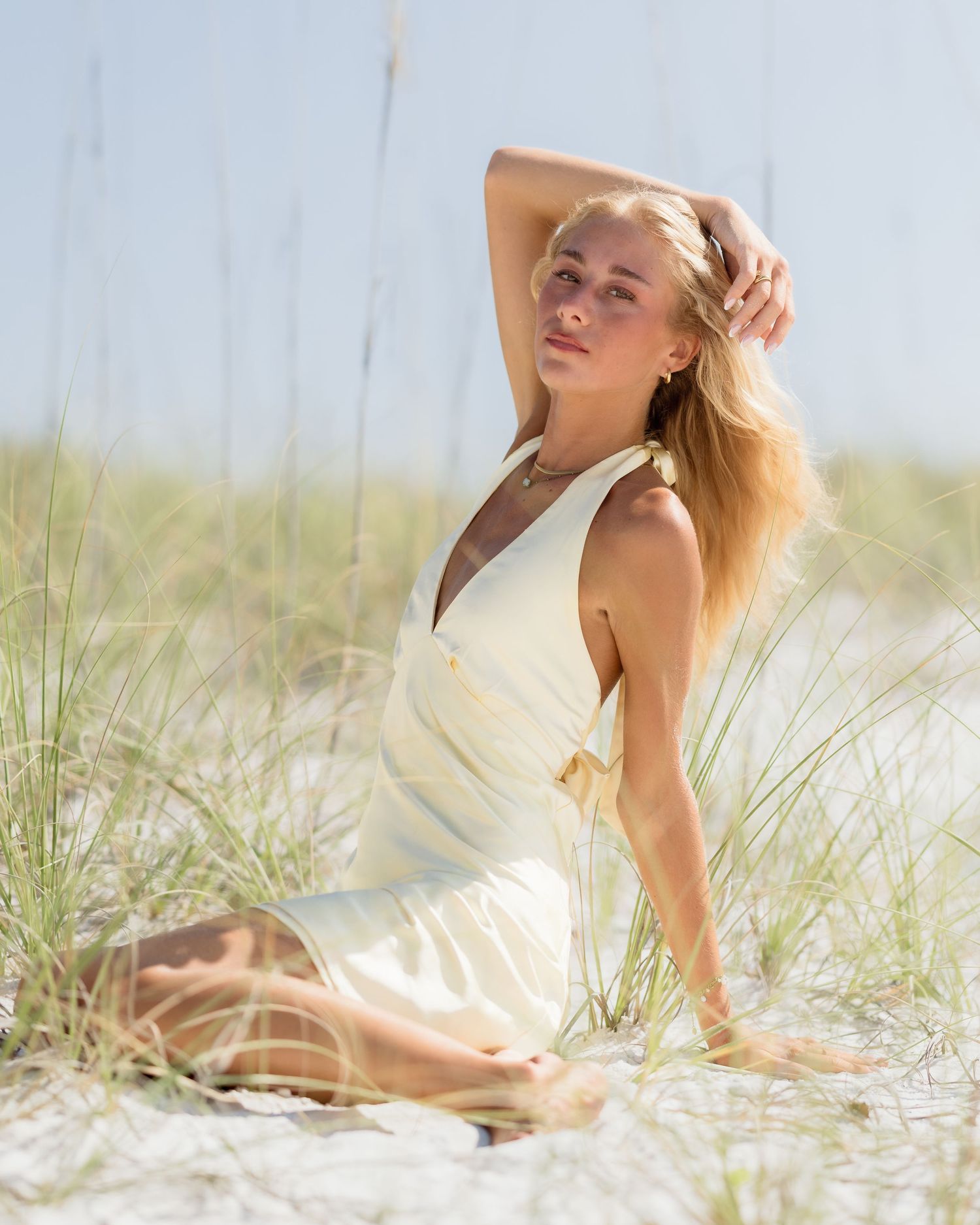 Blonde woman in white dress sitting on sandy beach dunes with sea grass in bright sunlight.