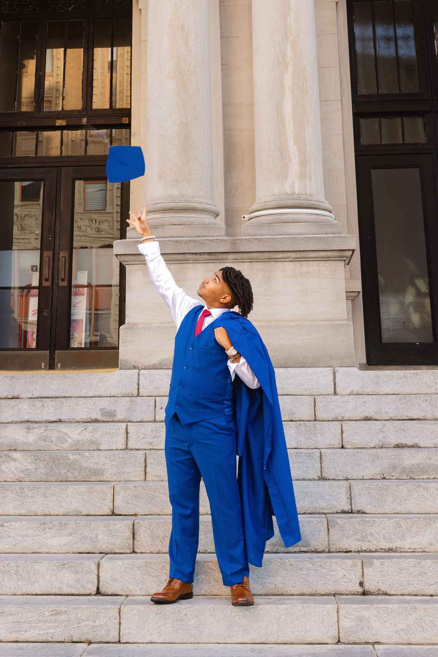 Graduate in blue cap and gown tosses mortarboard in the air on university steps, celebrating academic achievement.