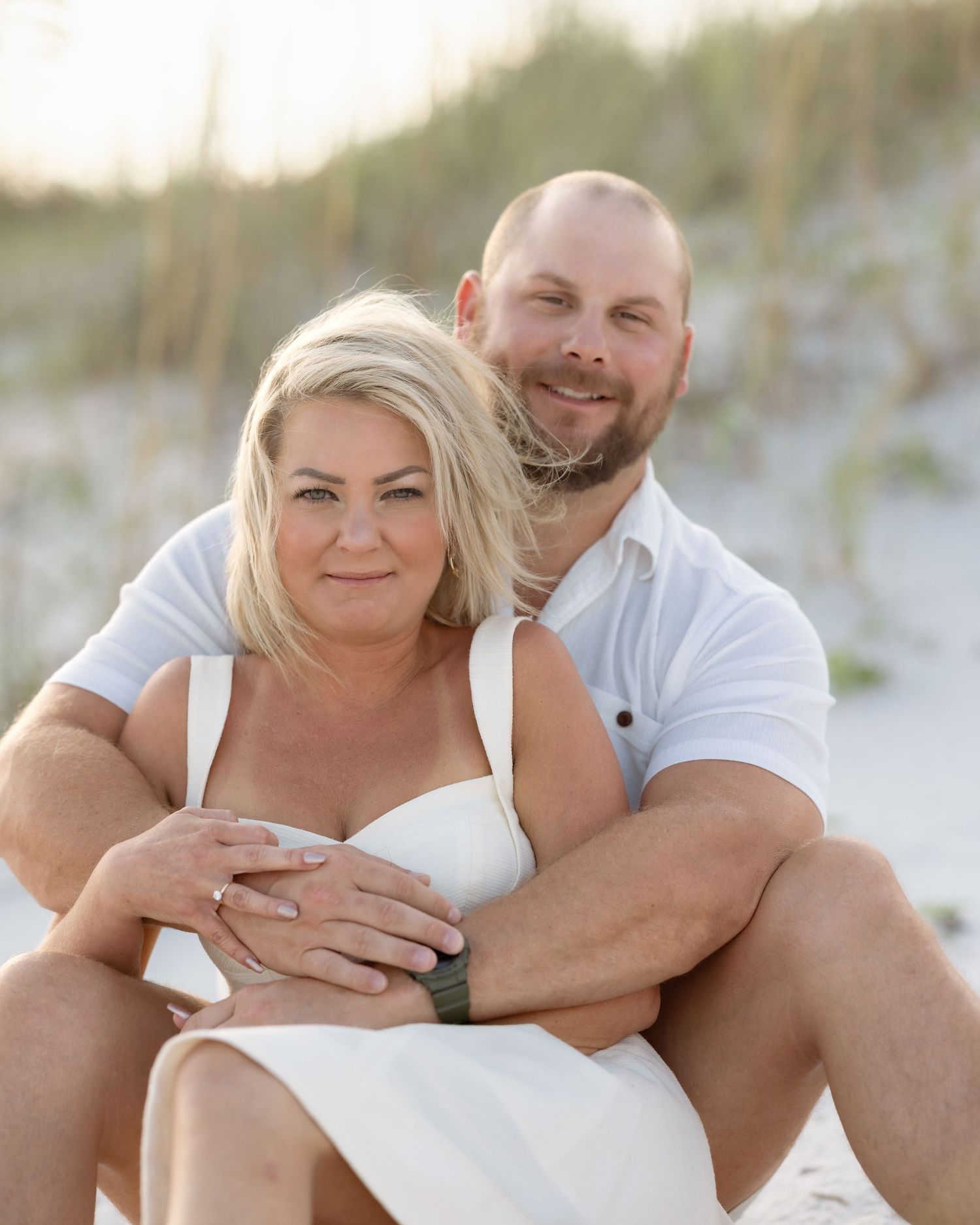 A smiling couple in white outfits embraces on a sandy beach with dunes and sea oats in the background.