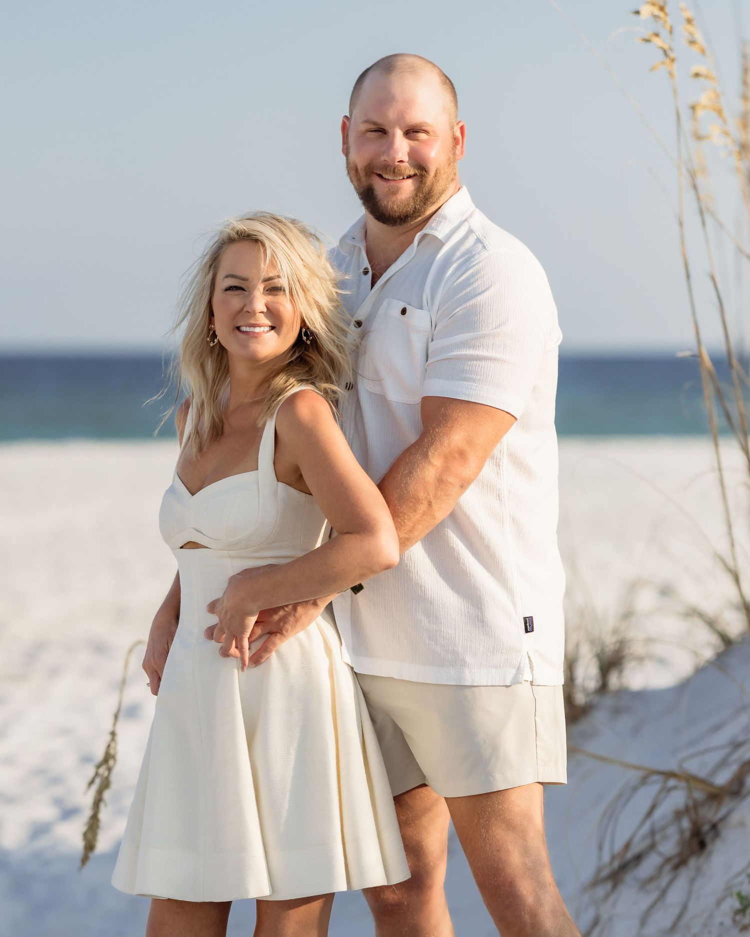 Smiling couple in white outfits posing together on a sunny white sand beach with sea oats in the background.