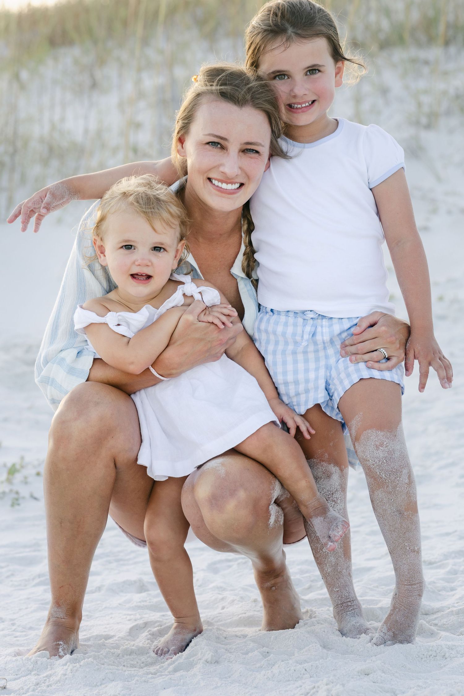 A smiling mother crouches on a sandy beach hugging her two young daughters, all barefoot and enjoying a sunny day.