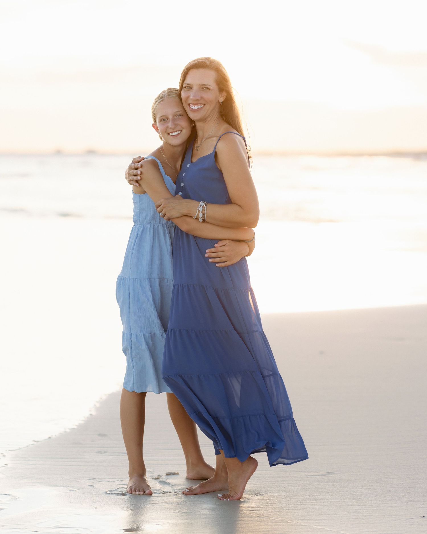 Two women in blue dresses embracing and smiling on a sandy beach at sunset.