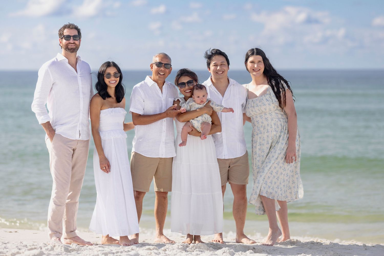 Multi-generational family posing together on a sunny beach, dressed in white and beige outfits, smiling with clear blue skies.