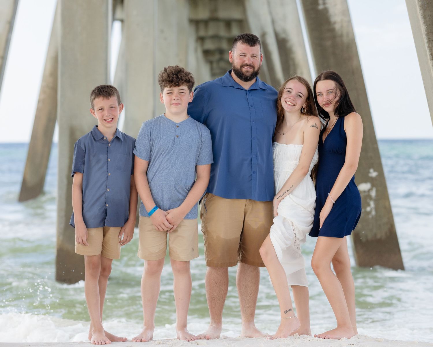 Family of five poses together under beach pier with ocean waves in the background.