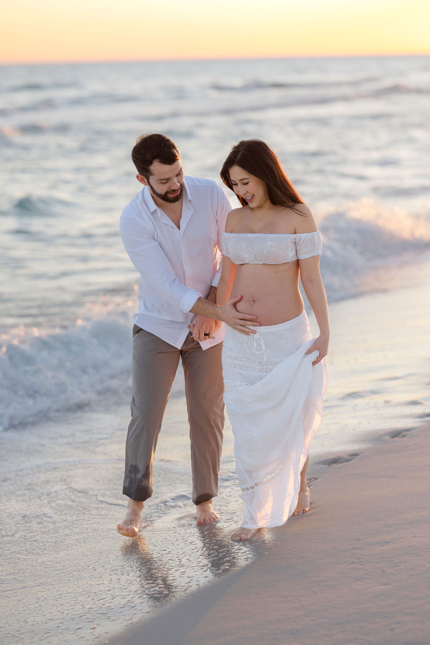 Pregnant couple walking barefoot on beach at sunset, man touching womans baby bump, wearing white outfits.