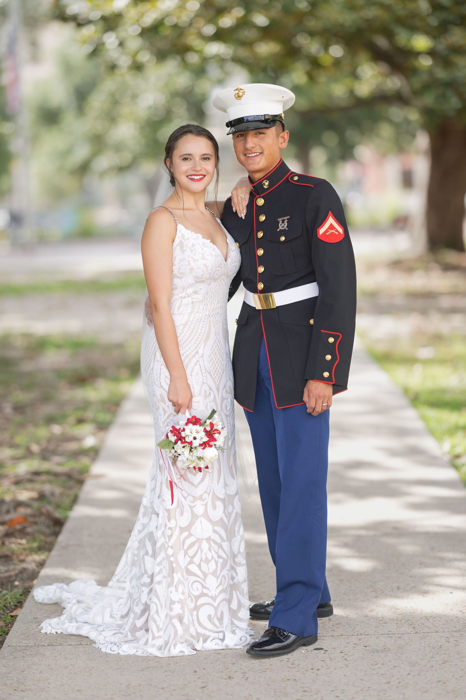 Marine in dress blues uniform poses with bride in white lace gown holding bouquet on outdoor pathway.