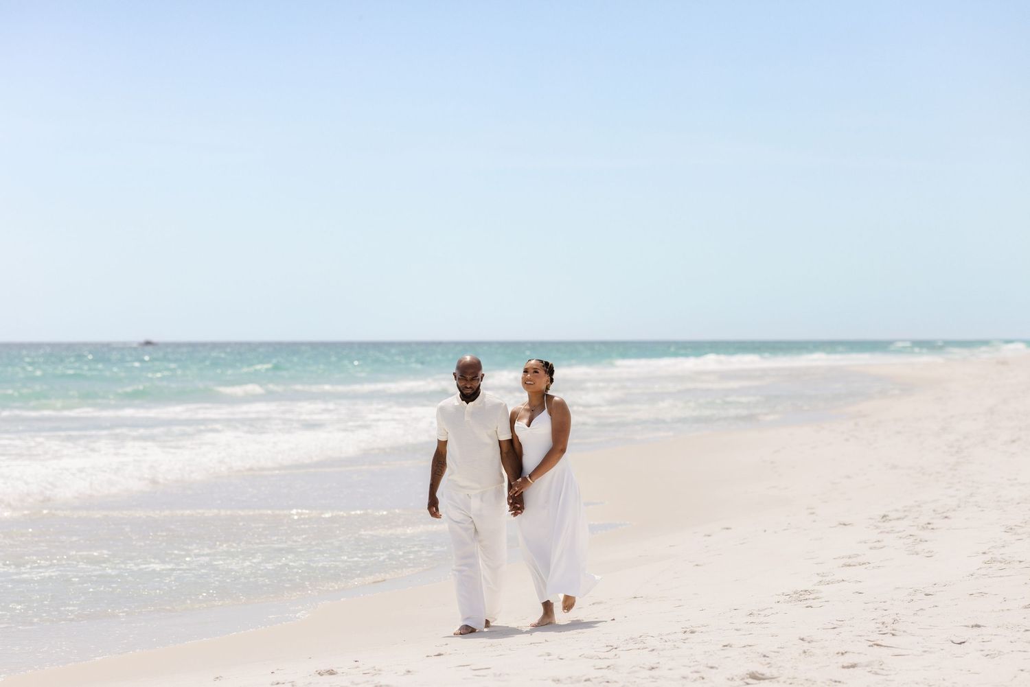Couple in white outfits walking hand-in-hand on a pristine white sandy beach with turquoise ocean waves.