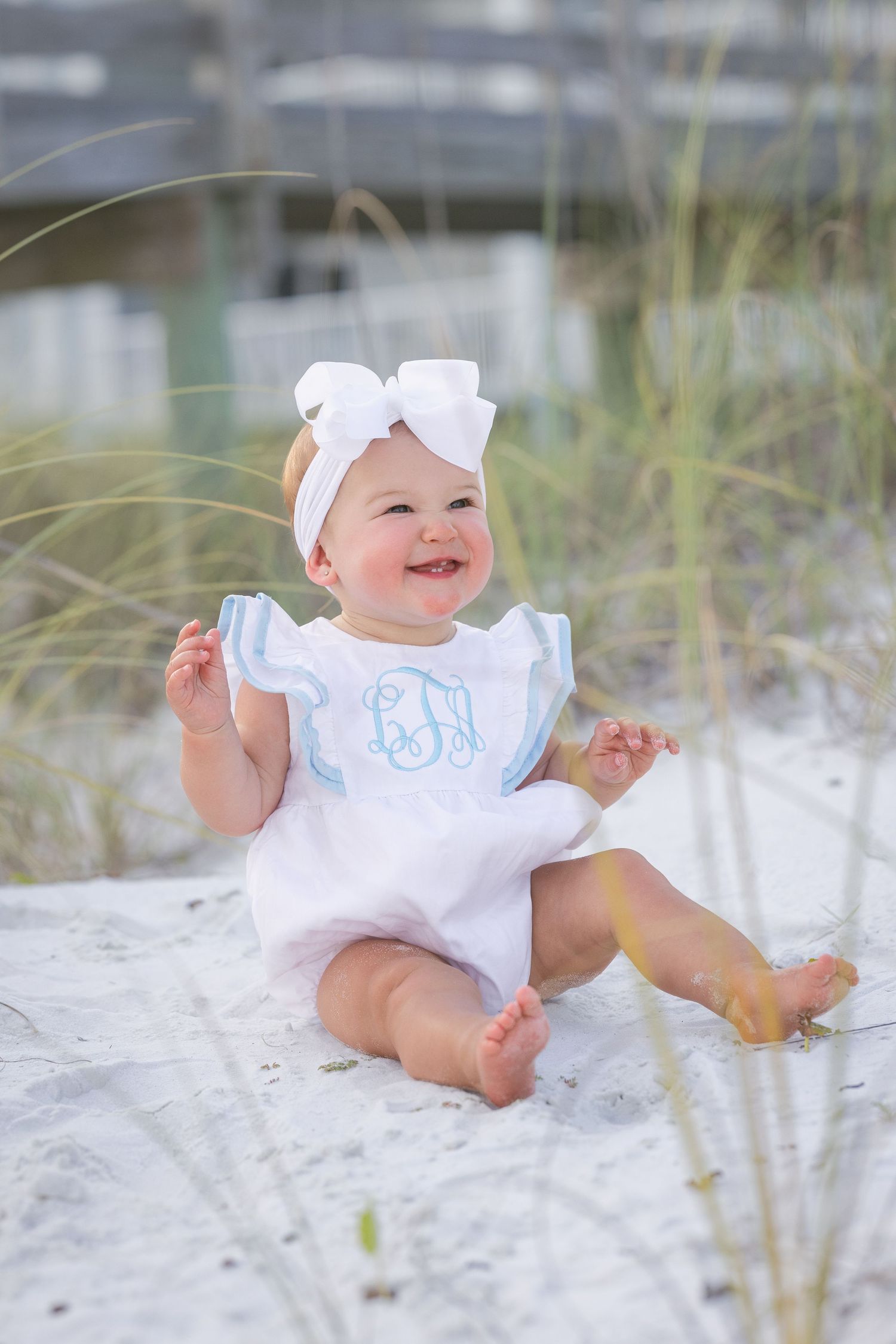 Happy baby girl in white monogrammed romper with bow headband sitting on white sand beach dunes smiling.