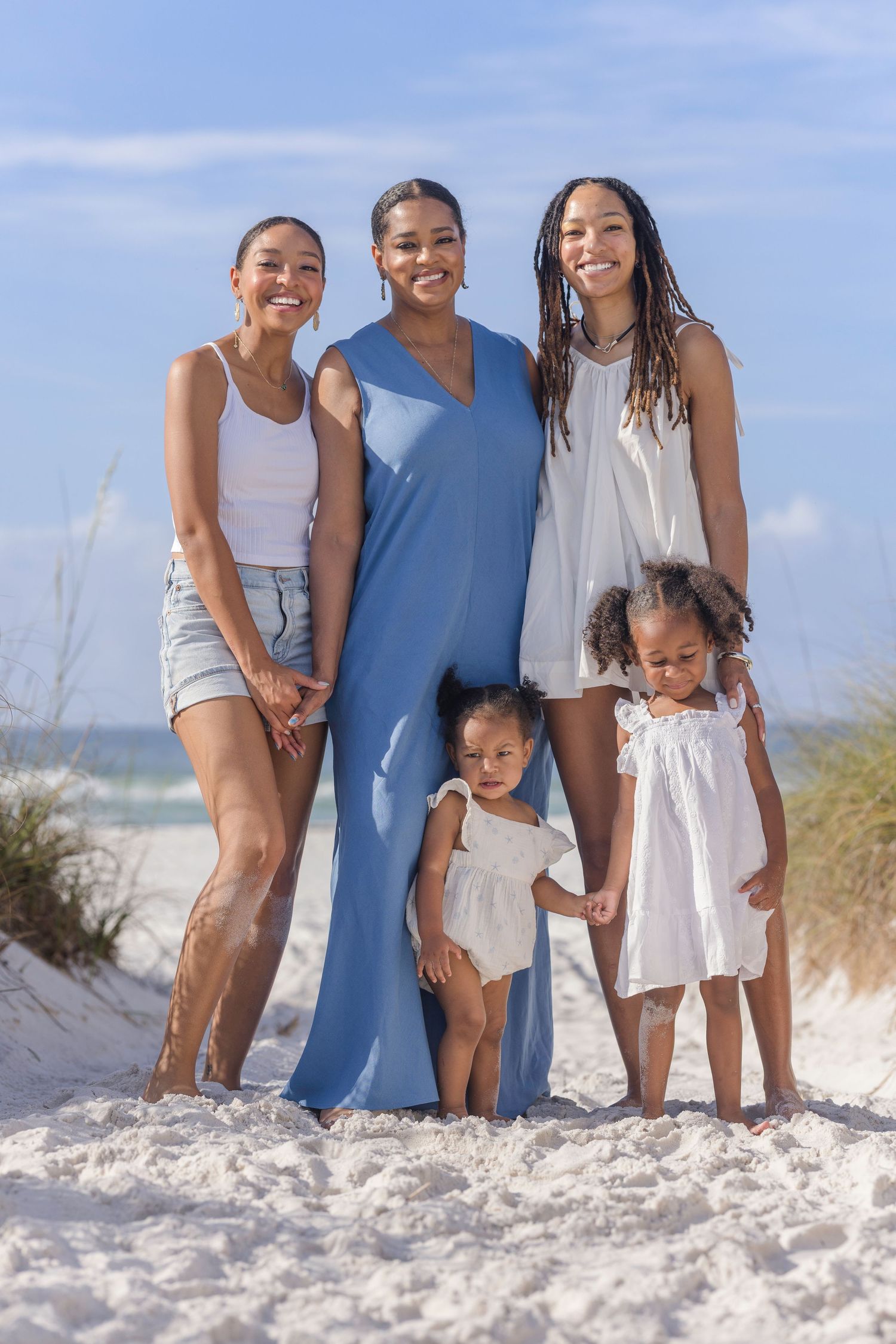 A joyful family of five poses together on a white sandy beach, with three women and two young girls smiling in summer outfits.