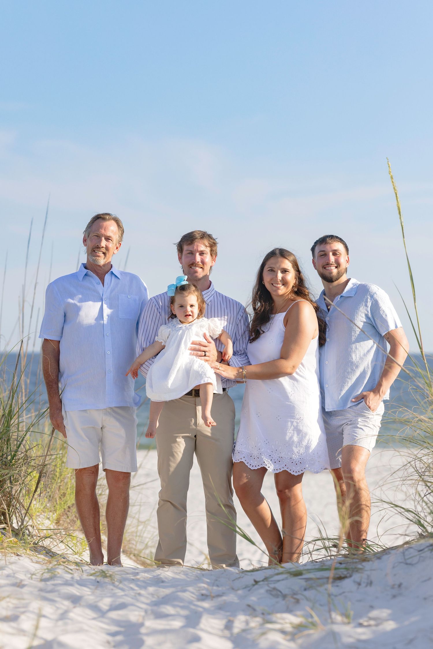 Family of five poses on sandy beach dunes with sea oats, dressed in white and blue, holding a baby girl.