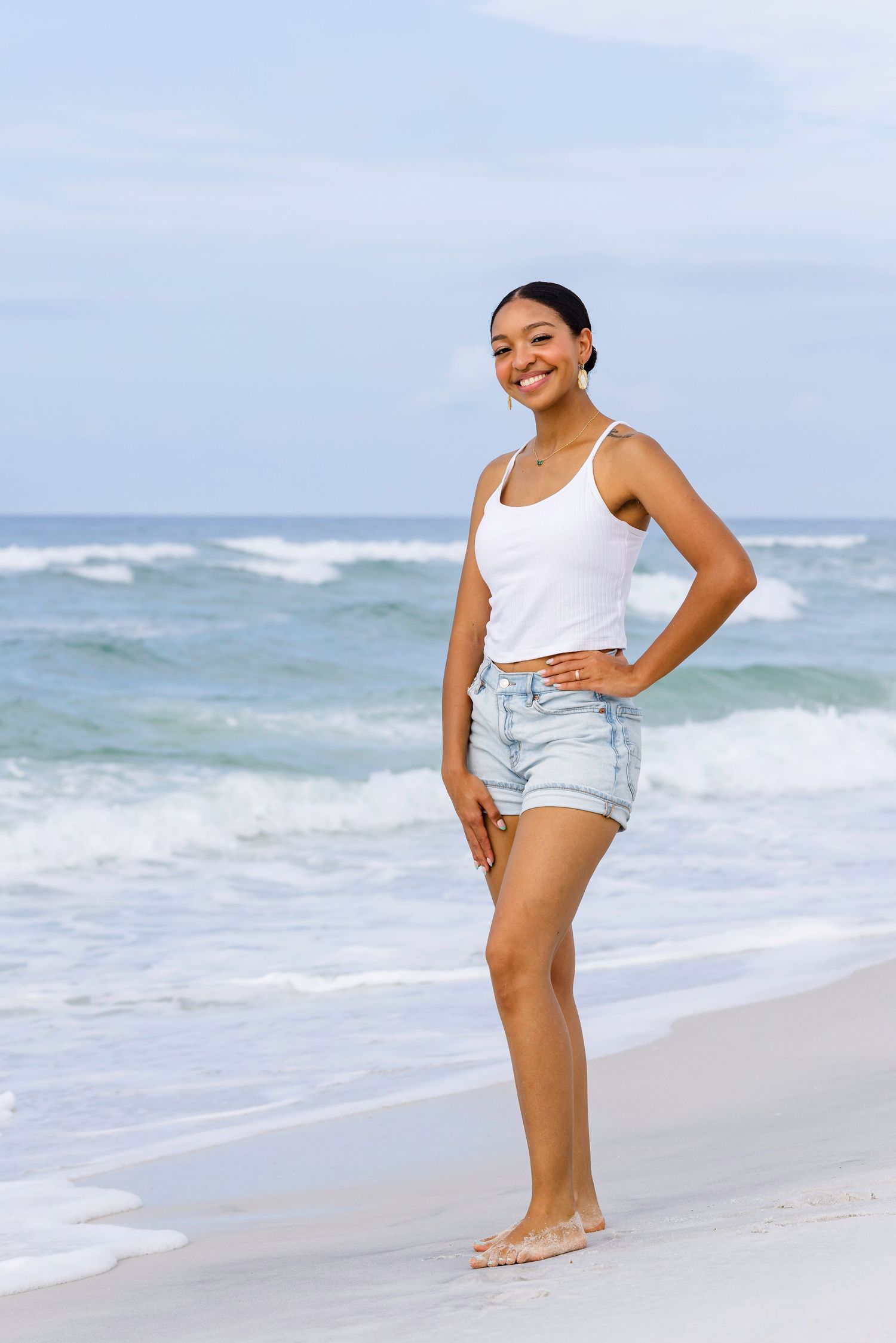 Smiling young woman in white tank top and denim shorts standing barefoot on a sunny beach with ocean waves.