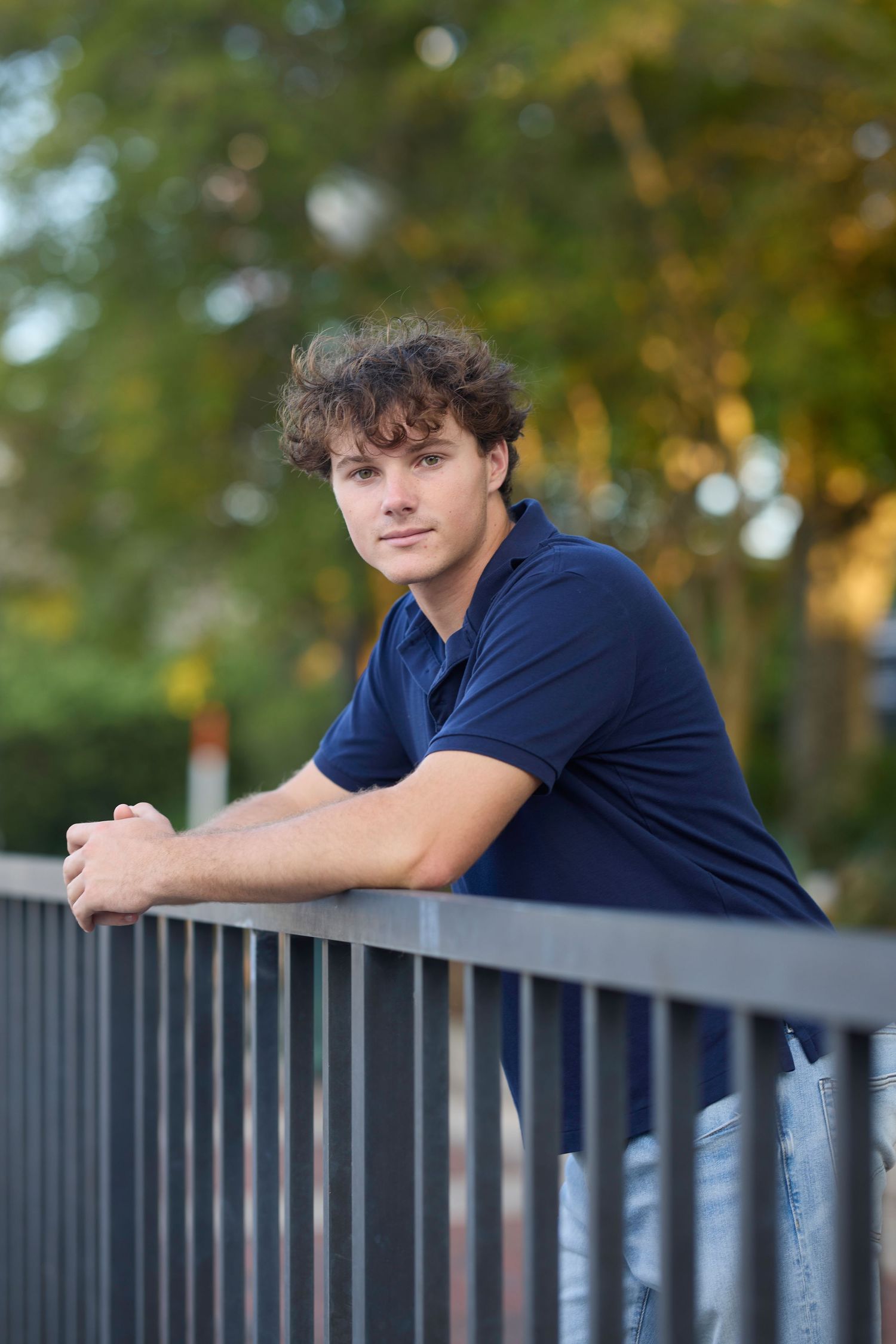 A figure leans against a dark metal railing while wearing a navy blue shirt on a sunny autumn day.