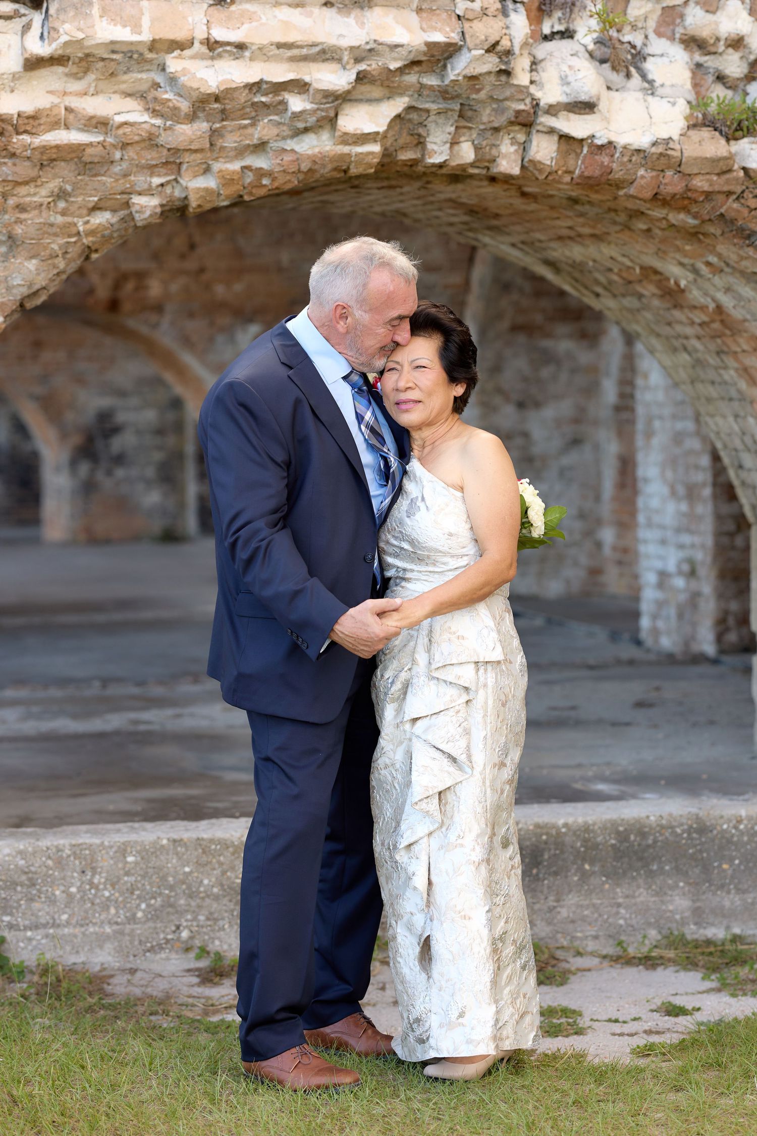 A romantic wedding couple embraces in front of stone archways at a historic fort or castle.