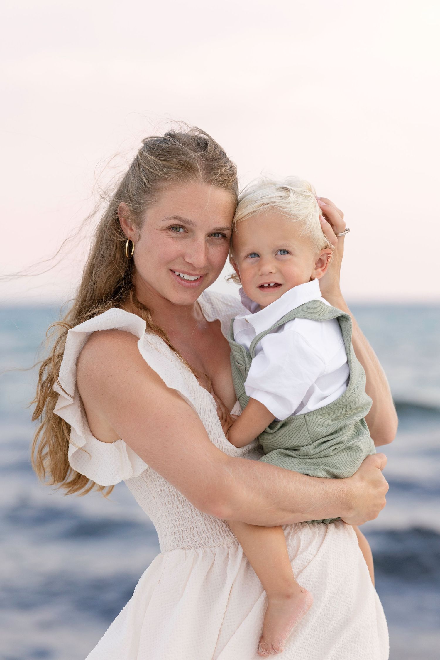 A mother and child share joyful moments together at the beach in white summer outfits.