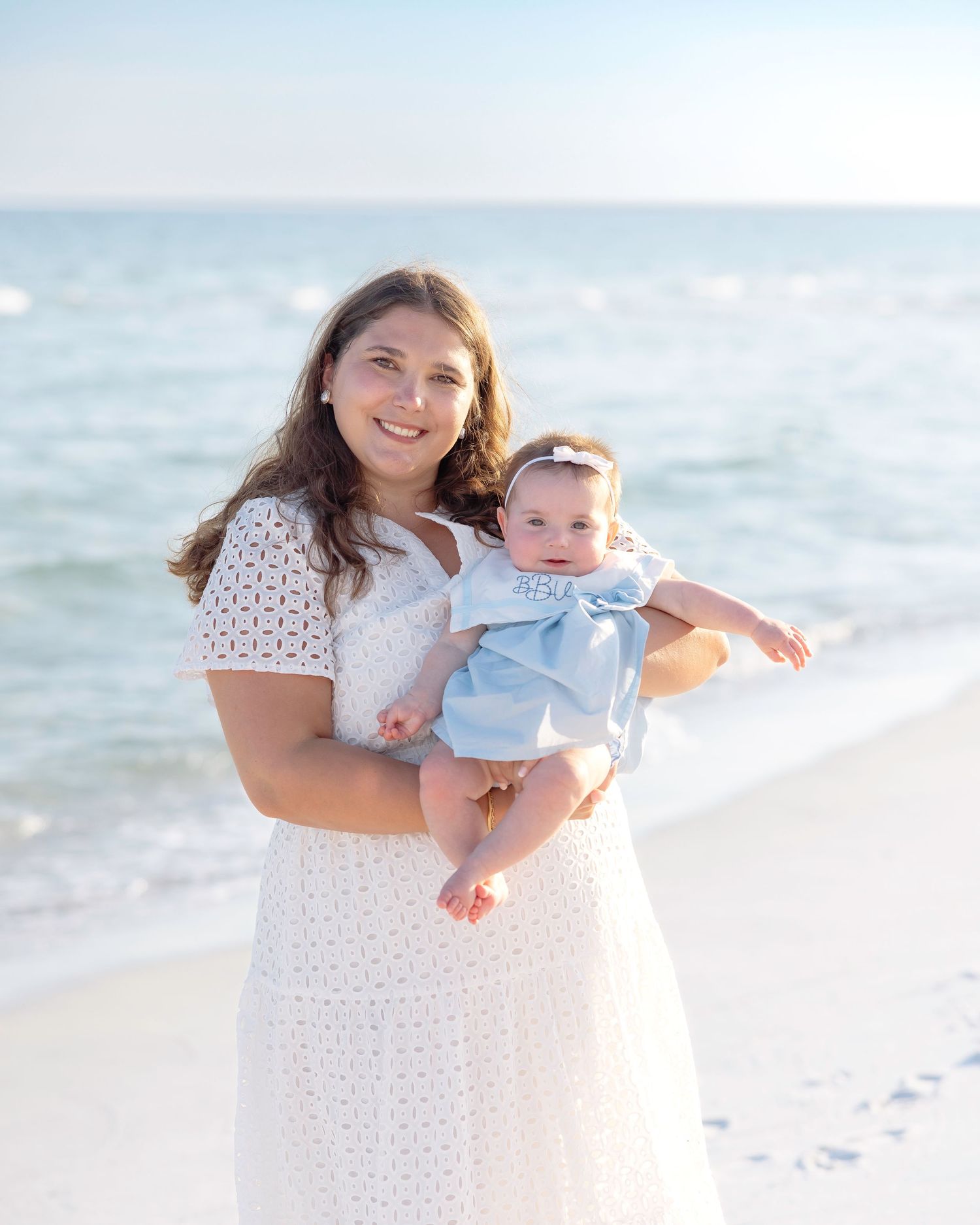 A mother and child share a joyful moment together on a sunny beach with gentle waves in the background.