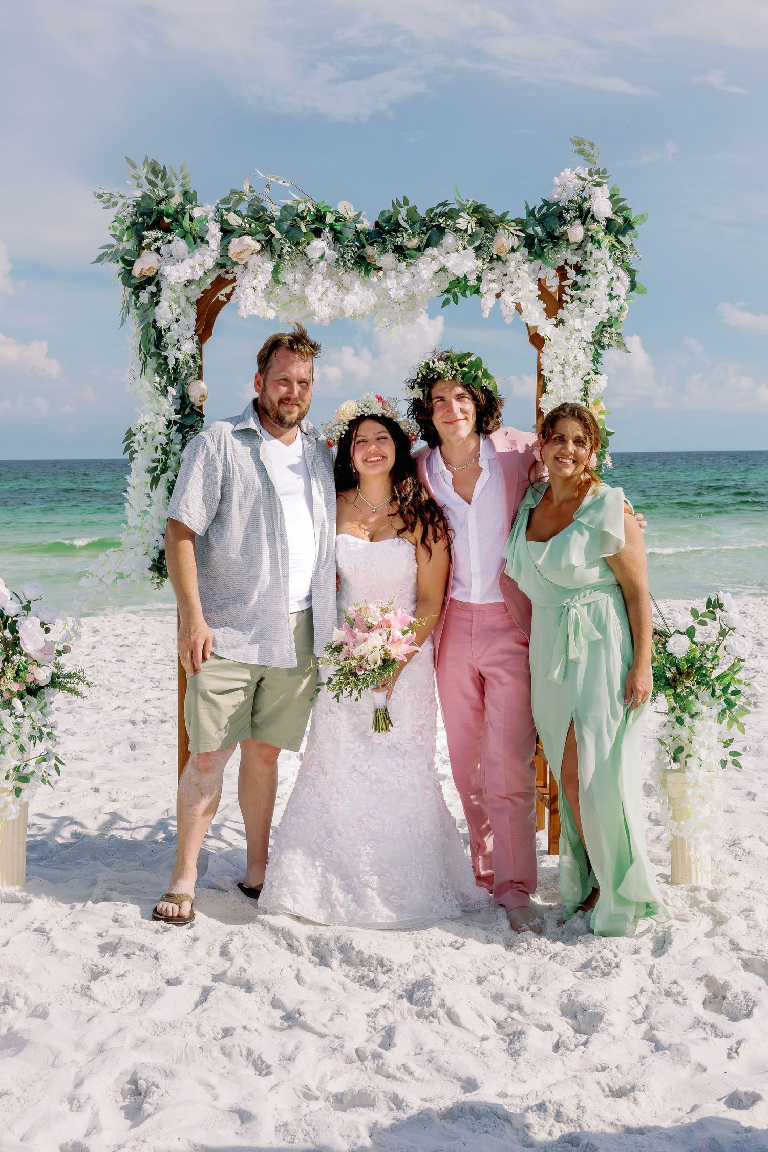 Beach wedding group photo under floral arch with ocean backdrop.