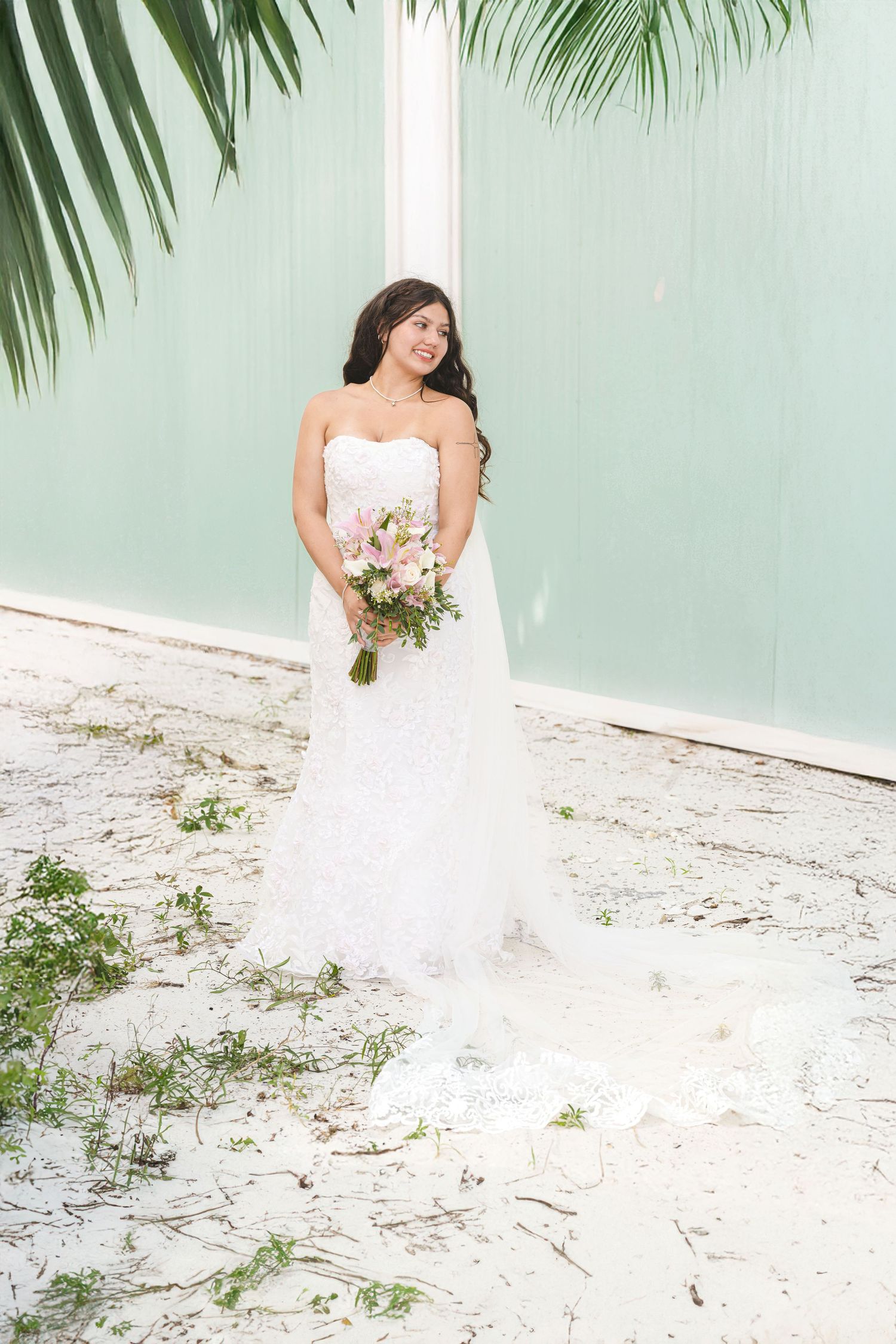 Bride in white wedding dress posing against mint green wall with palm fronds.