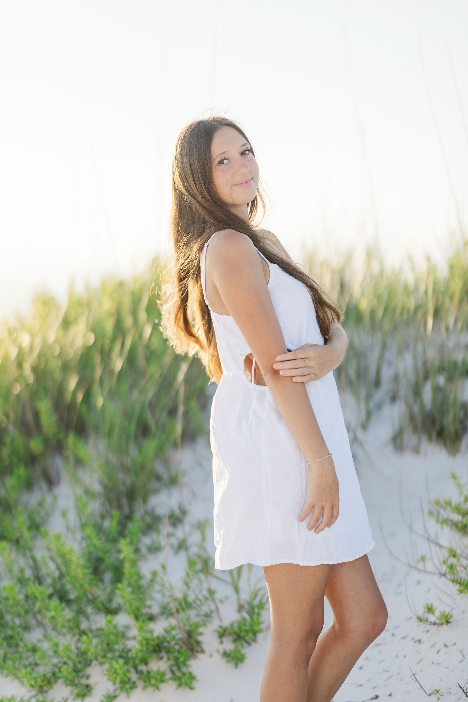 Person in white summer dress poses on sand dunes with beach grass in background.