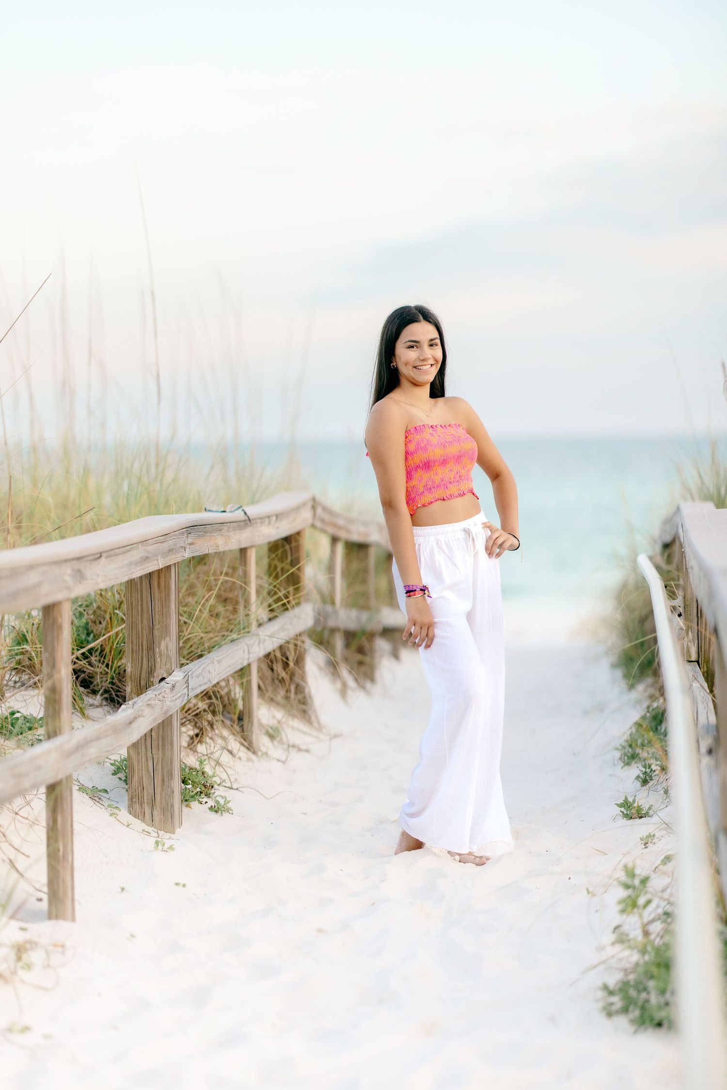 Beach boardwalk portrait with coral top and white pants against coastal scenery.