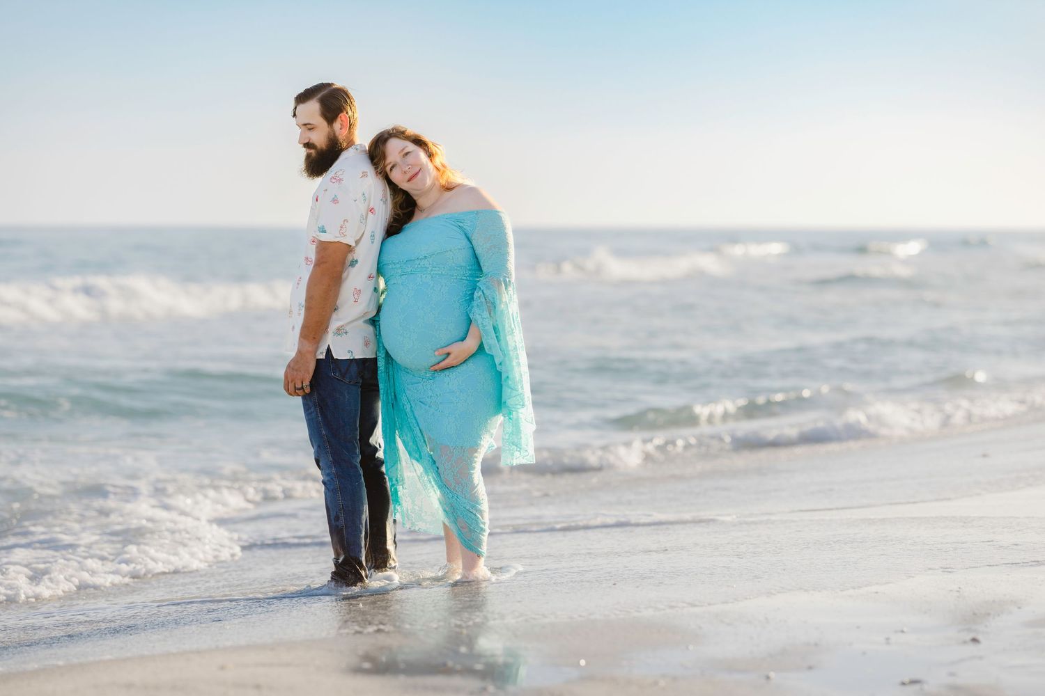 Pregnant couple standing on a beach, woman in teal maternity gown holding her belly, man in white shirt and jeans beside her.