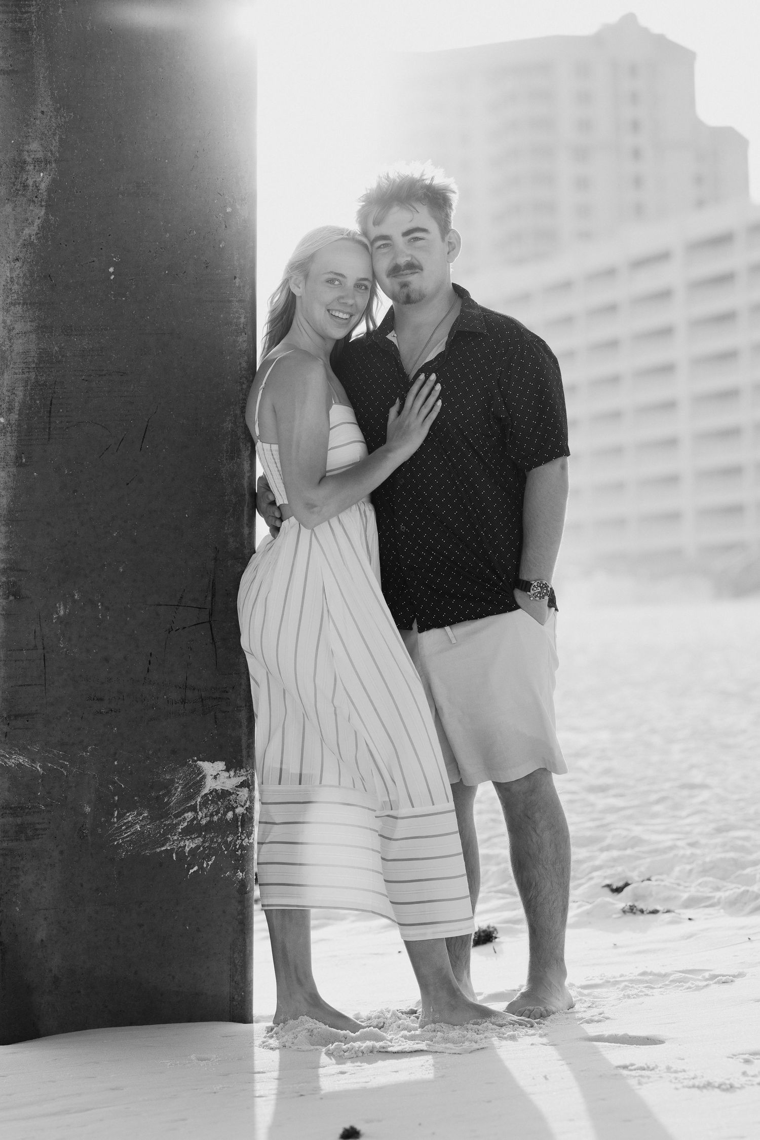 Black and white photo of a couple embracing near a beach pier, woman in striped dress smiling at camera.