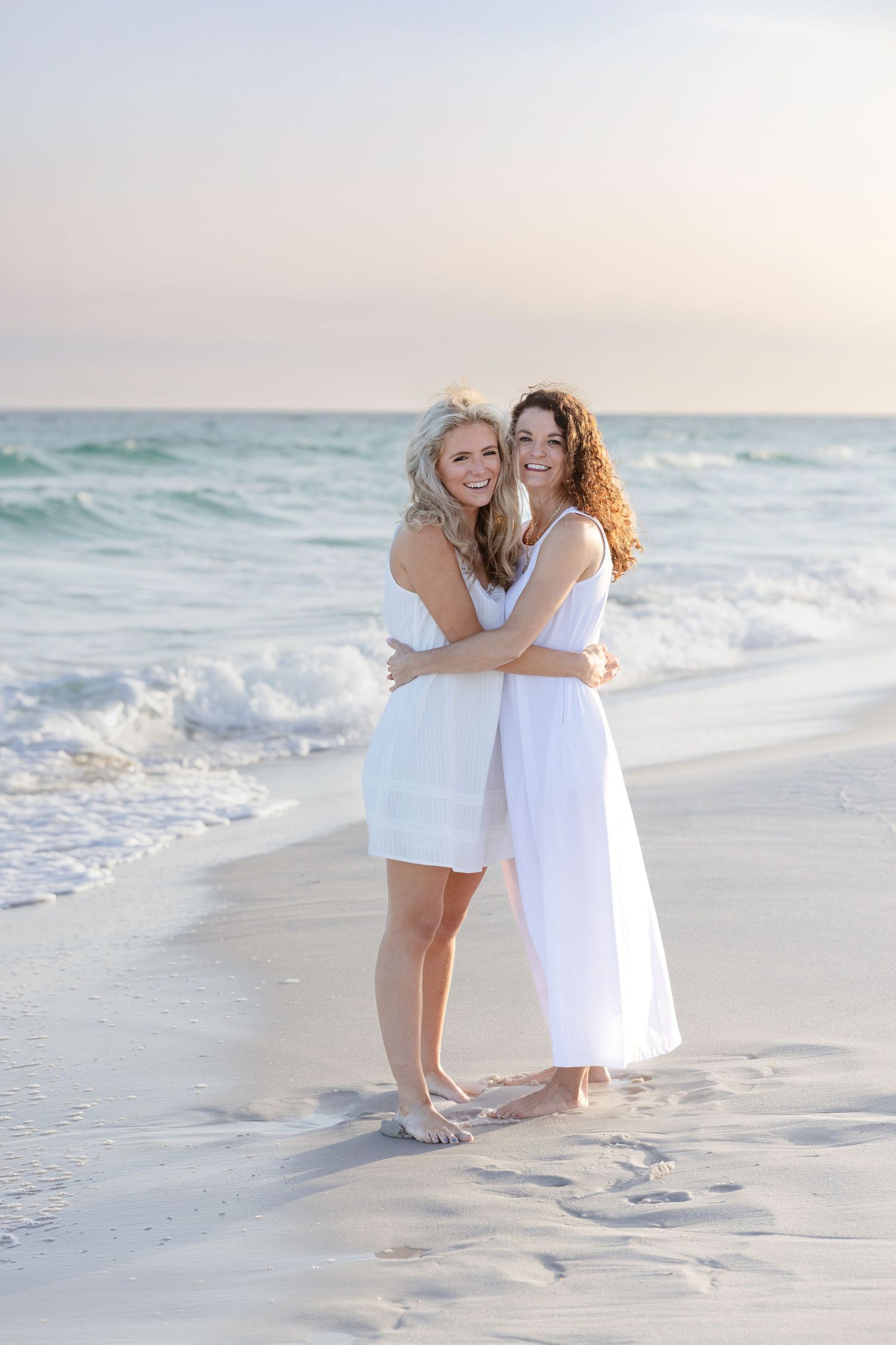 Two women in white dresses embracing on a sandy beach at sunset with ocean waves in the background.