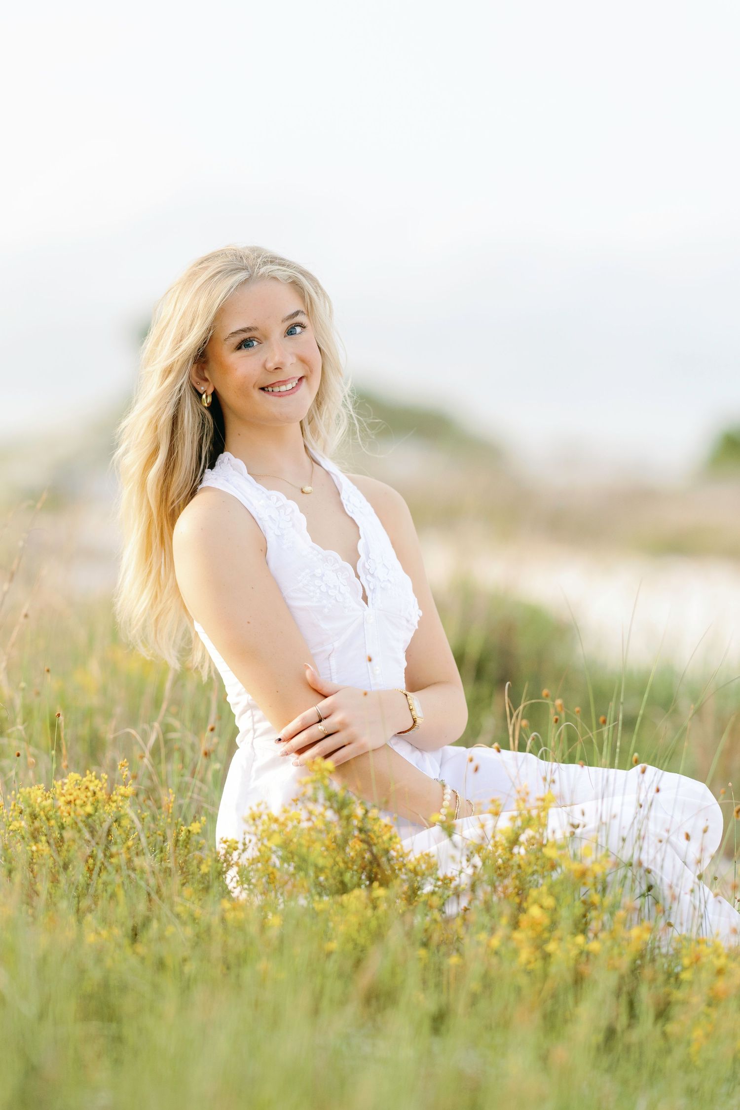 A person in a white sleeveless top sits among blooming yellow wildflowers in a sunny meadow.
