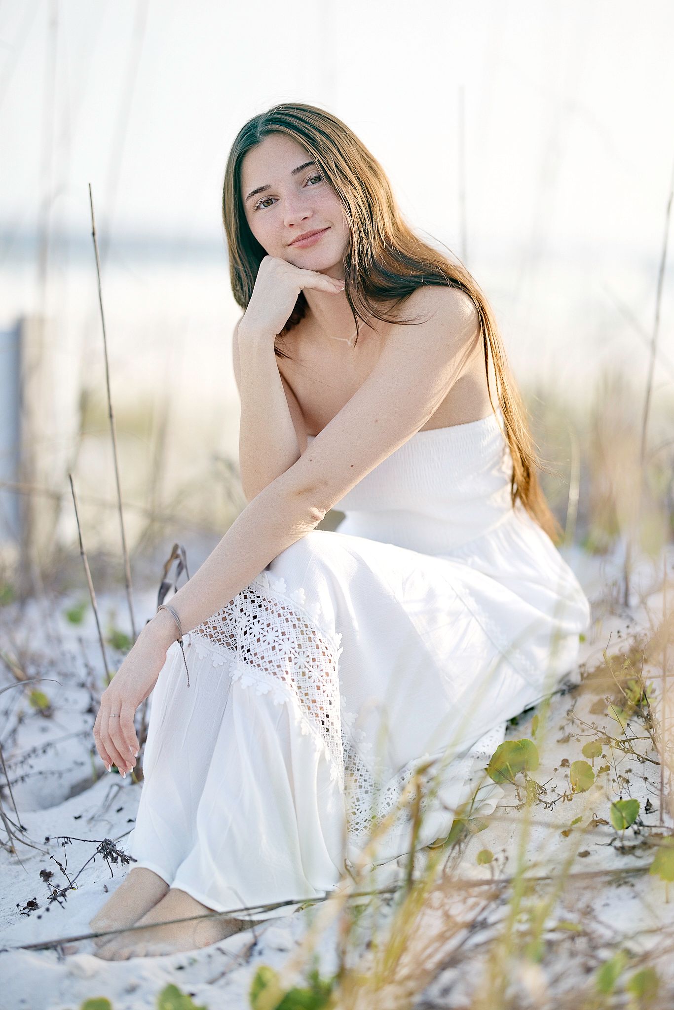 Ethereal portrait in a flowing white dress amid coastal grasses and sand dunes at sunset.