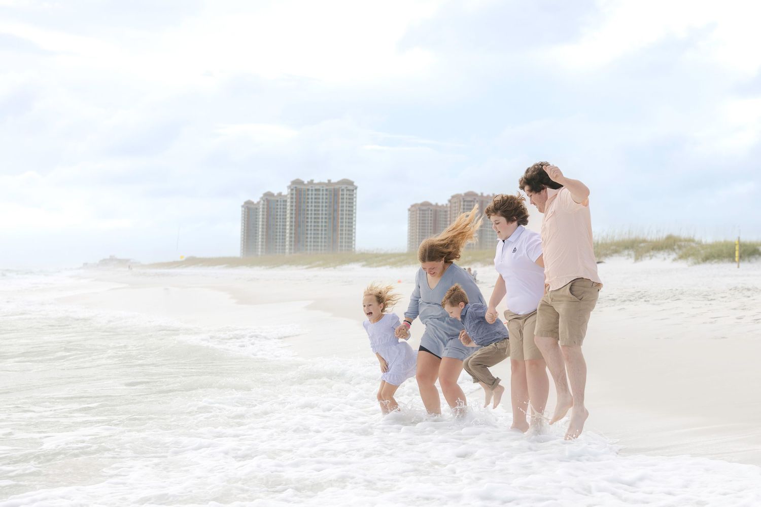 Family walking together and playing on a sunny beach with ocean waves and high-rise buildings in the background.